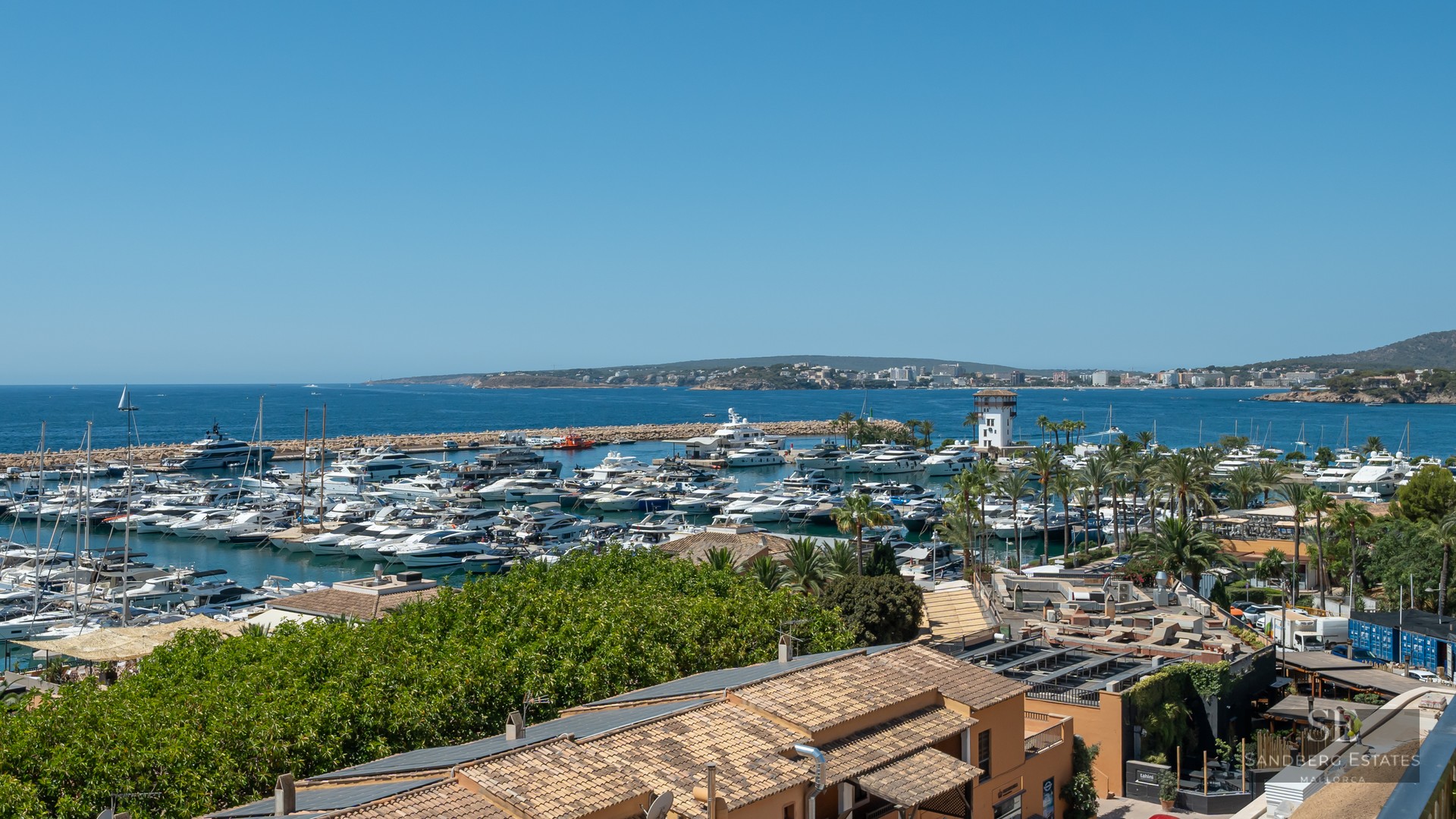 Panoramic view of a luxury marina filled with white yachts under a clear blue sky, overlooking the Mediterranean Sea.