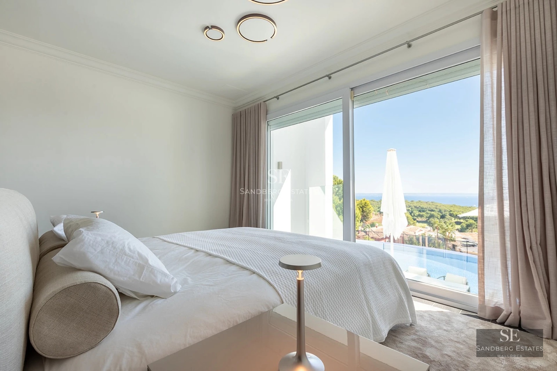 Bright master bedroom with a white bed and floor-to-ceiling glass doors overlooking a pool and the sea.