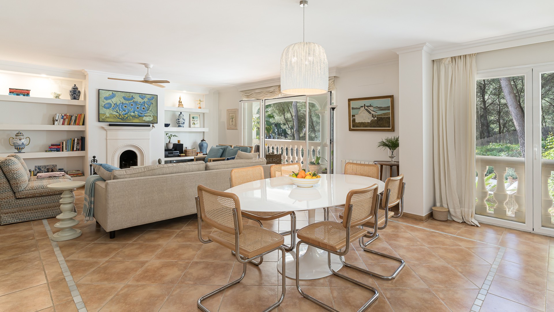 Open-plan living and dining room with terracotta tiles, white oval table, cane chairs, and views of pine trees.
