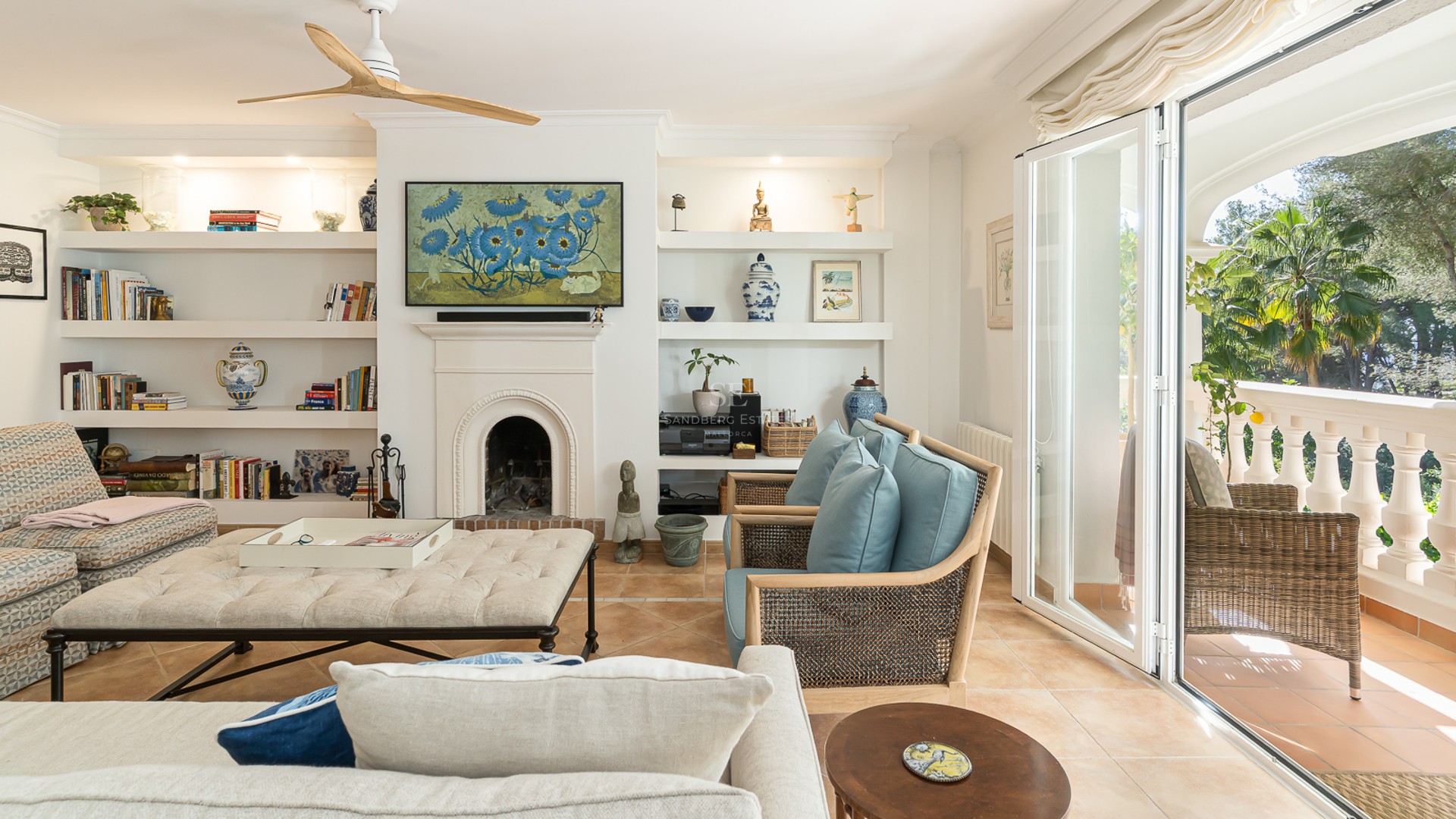 Bright living room with terracotta floors, white fireplace, built-in shelves, and glass doors opening to a lush terrace.