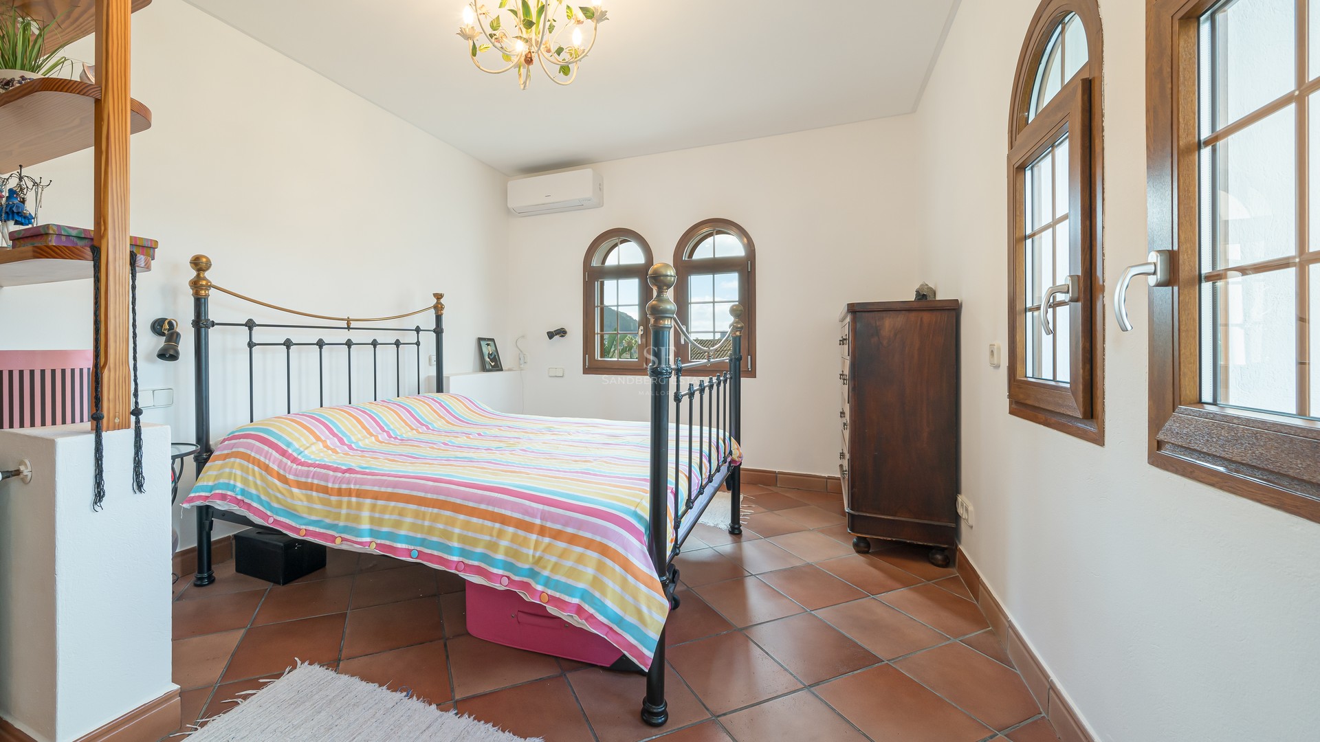 Bright bedroom featuring terracotta tile flooring, a wrought iron bed frame, and traditional arched wooden windows.