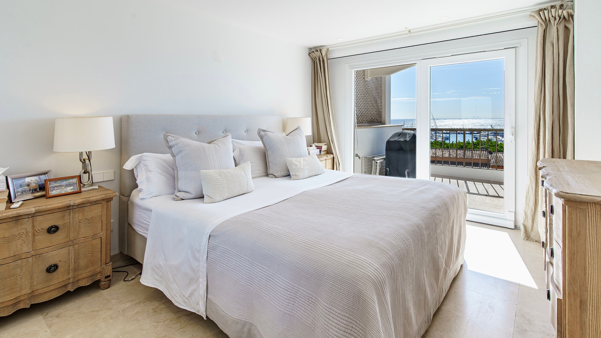 Bright bedroom featuring a large bed, wooden nightstands, and sliding glass doors with views of the sea and marina.