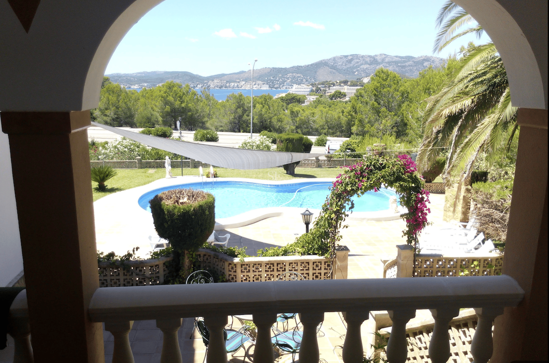 Vista desde un arco de una piscina turquesa, jardín frondoso y el mar Mediterráneo al fondo.