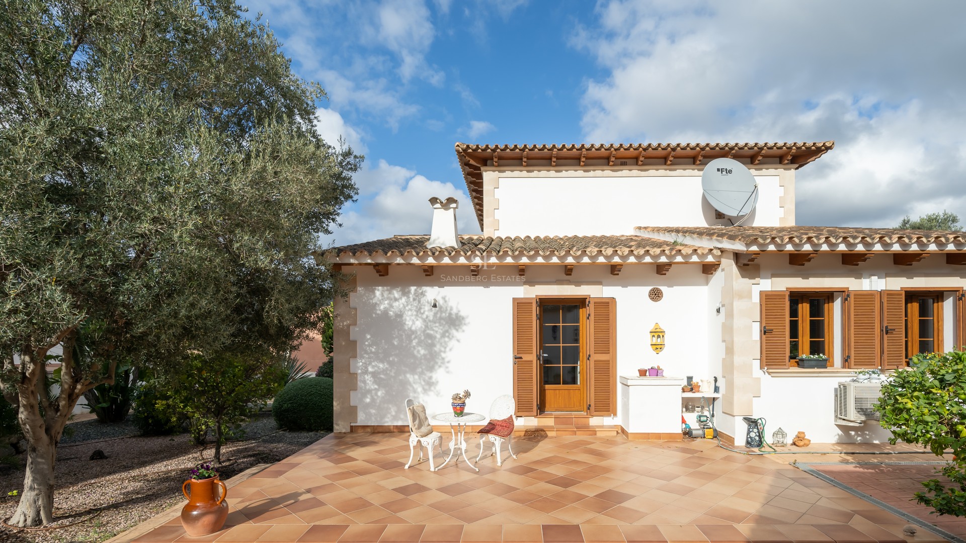 Villa terrace with terracotta tiles, wooden shutters, white outdoor seating, and a large olive tree under a blue sky.