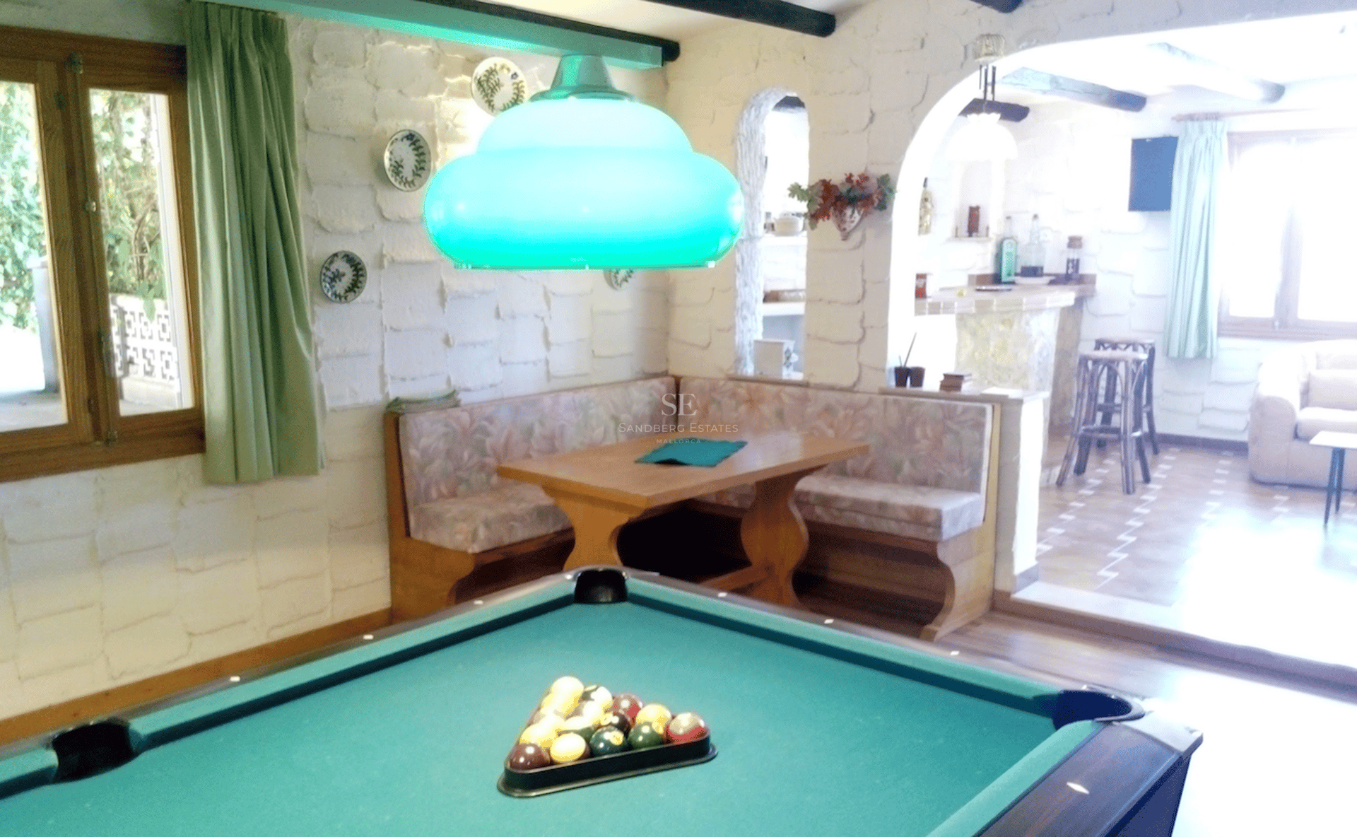 Sala de juegos rústica con mesa de billar, rincón de asientos de madera y paredes de piedra blanca.