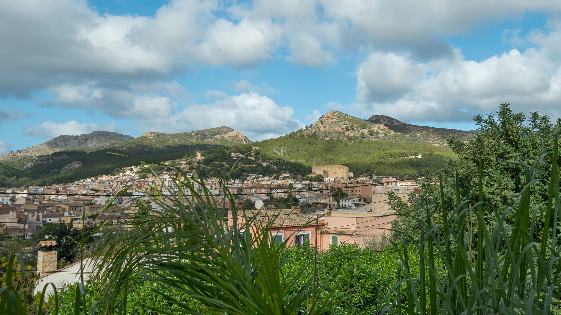 A scenic view of a Mediterranean town with terracotta roofs nestled against lush green mountains under a cloudy blue sky.