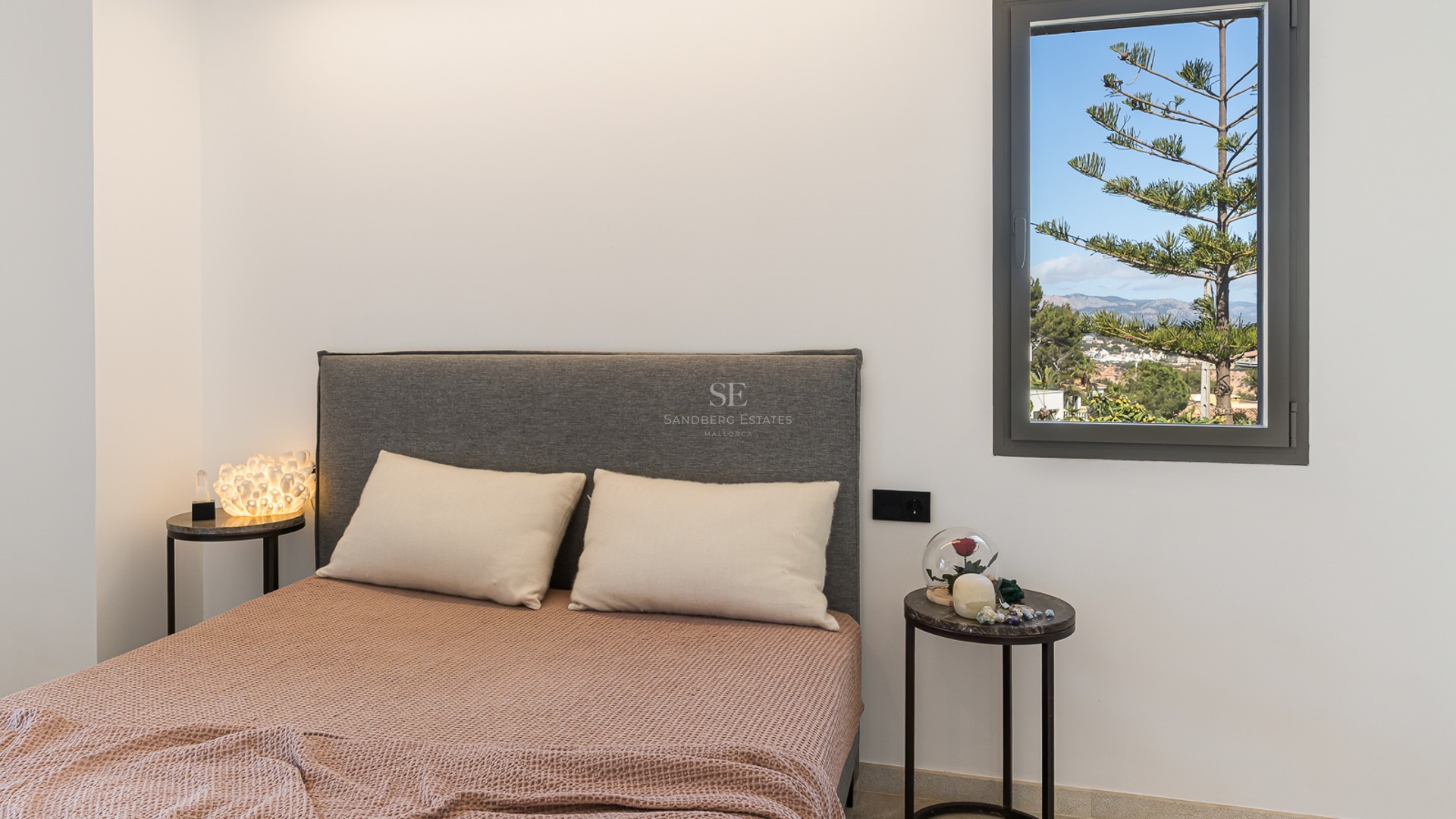 Säng med grå gavel, terrakottafärgat överkast och marmorbord framför ett fönster med utsikt över berg och träd.