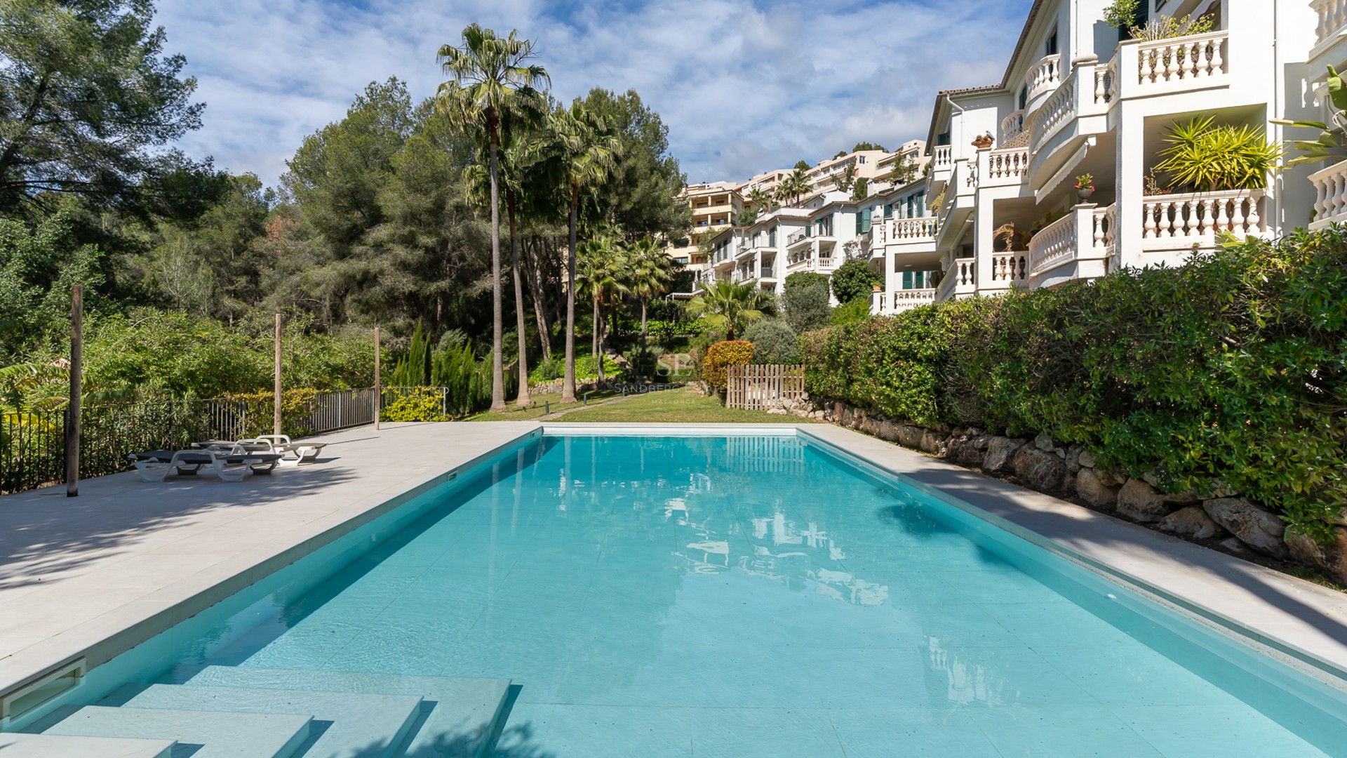 Large rectangular turquoise swimming pool with white stone decking, surrounded by palm trees and white apartments.