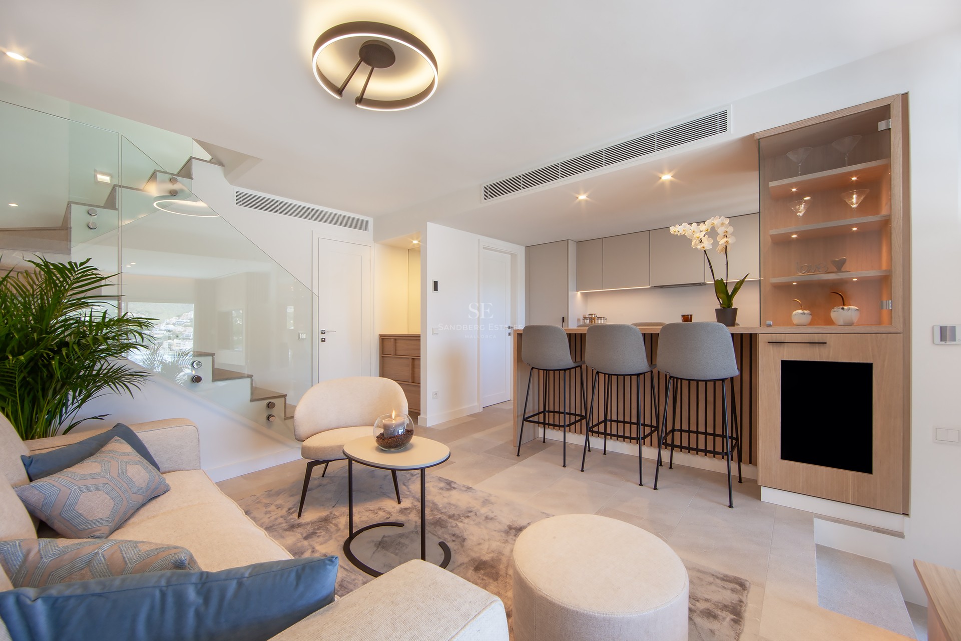 Salon moderne avec canapé crème, escalier en verre et bar à petit-déjeuner en bois avec tabourets gris.