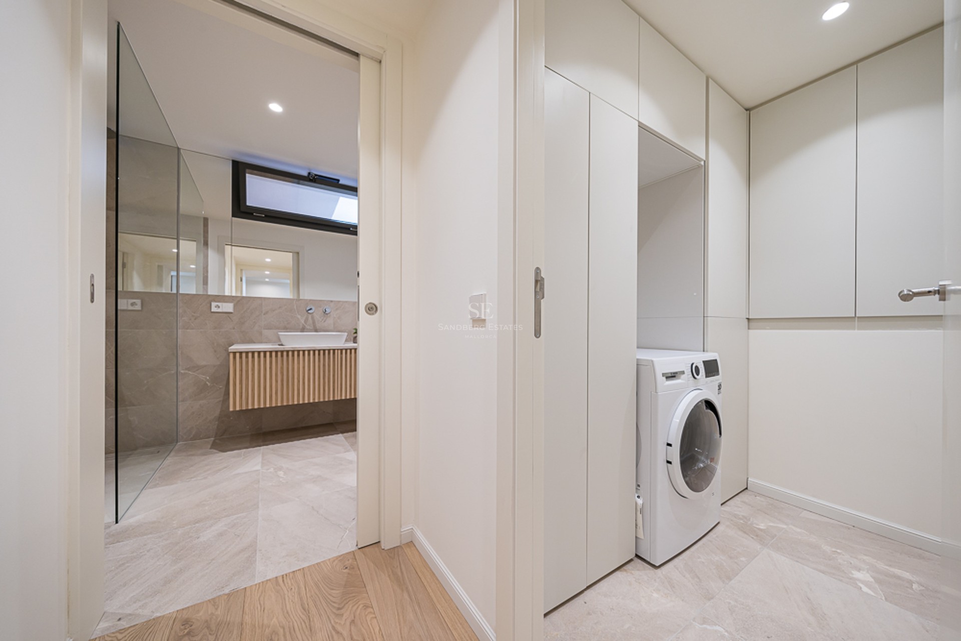A white laundry cupboard with integrated washing machine next to a modern bathroom with marble tiles and wood vanity.