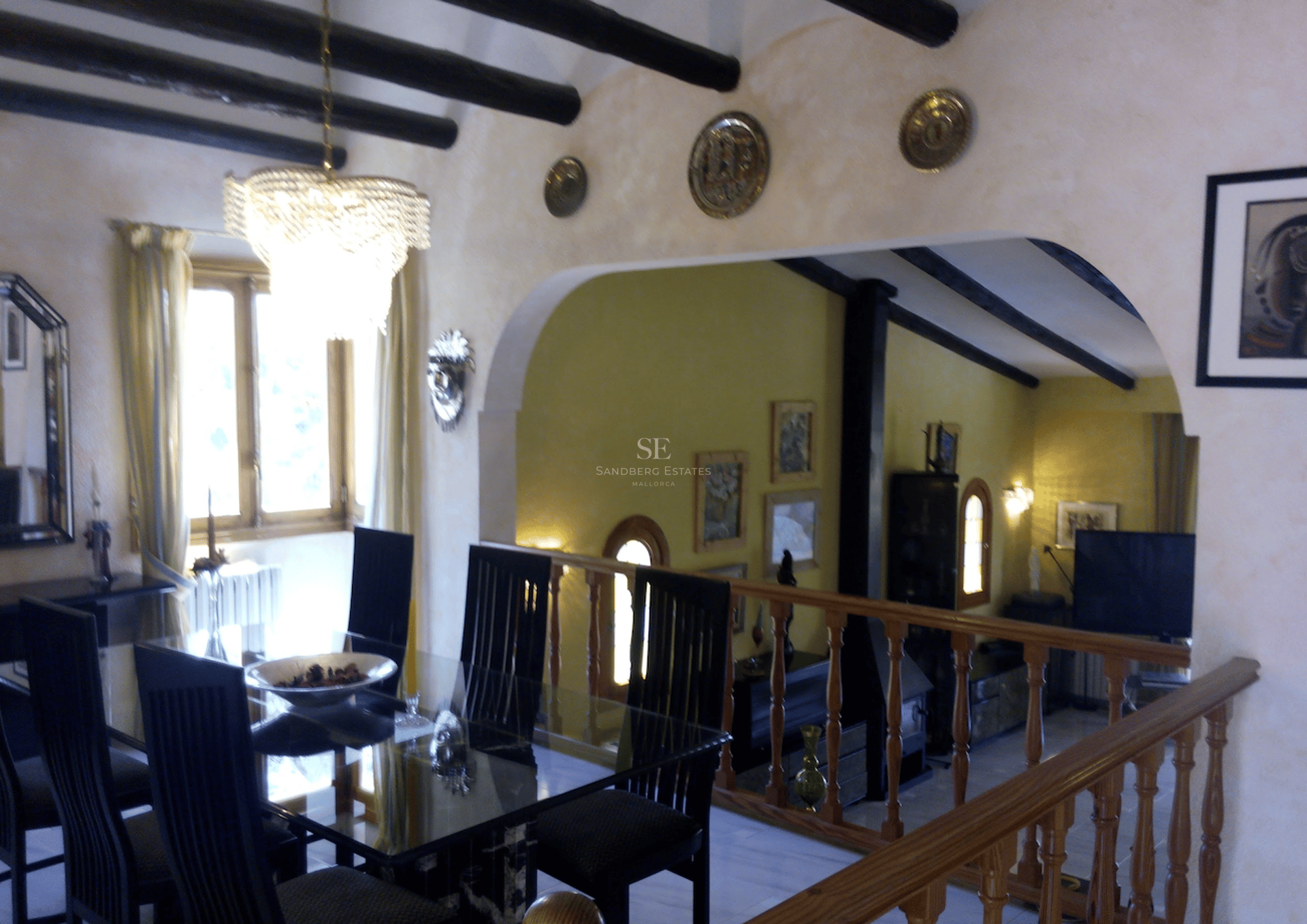 Comedor con mesa de cristal y vigas de madera oscura, con balustrada de madera con vistas a un salón en el nivel inferior.