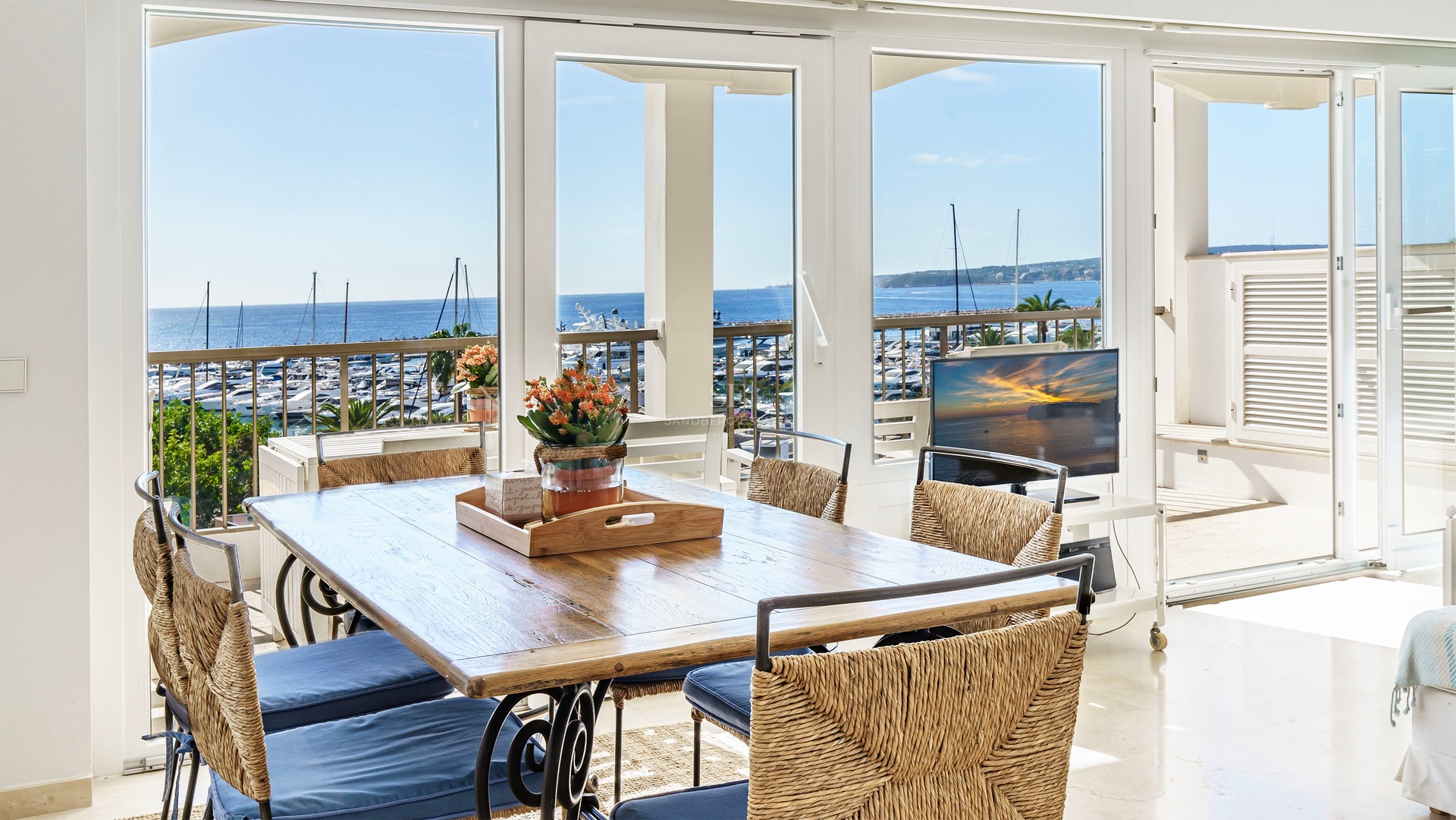 Sunlit dining room with a wooden table and woven chairs looking out over a yacht-filled marina and the blue ocean.