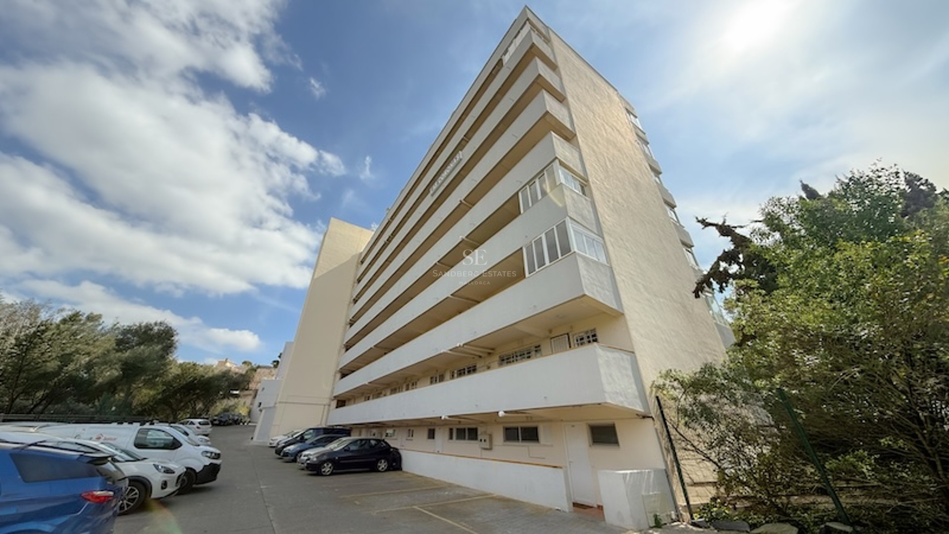 Vista exterior de un edificio de apartamentos blanco de varias plantas con amplios balcones y zona de aparcamiento.