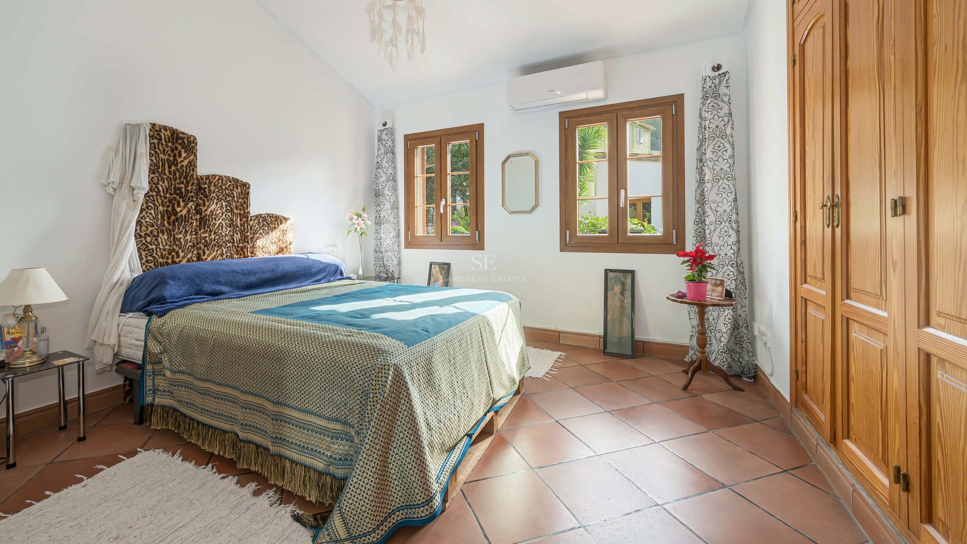Sunny bedroom with terracotta tile floors, large wooden built-in wardrobes, and windows overlooking greenery.