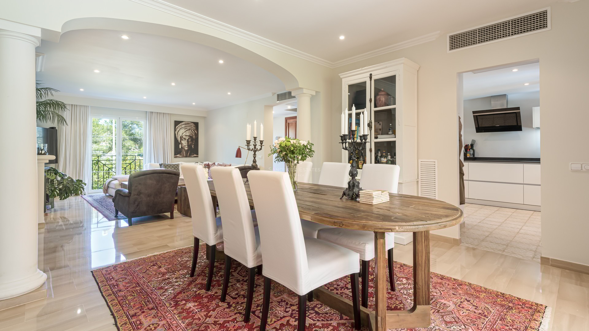 A rustic wooden dining table with white chairs on a red patterned rug, overlooking a living area and modern kitchen.