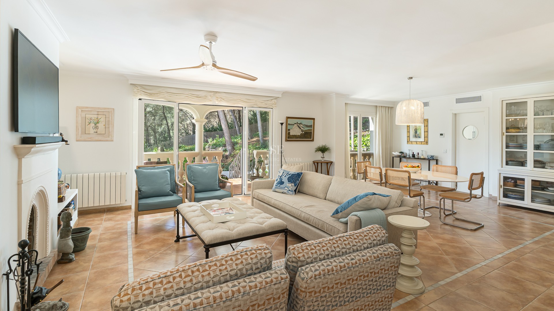 Spacious living room with terracotta floors, cream sofas, fireplace, and glass doors opening to a lush garden.