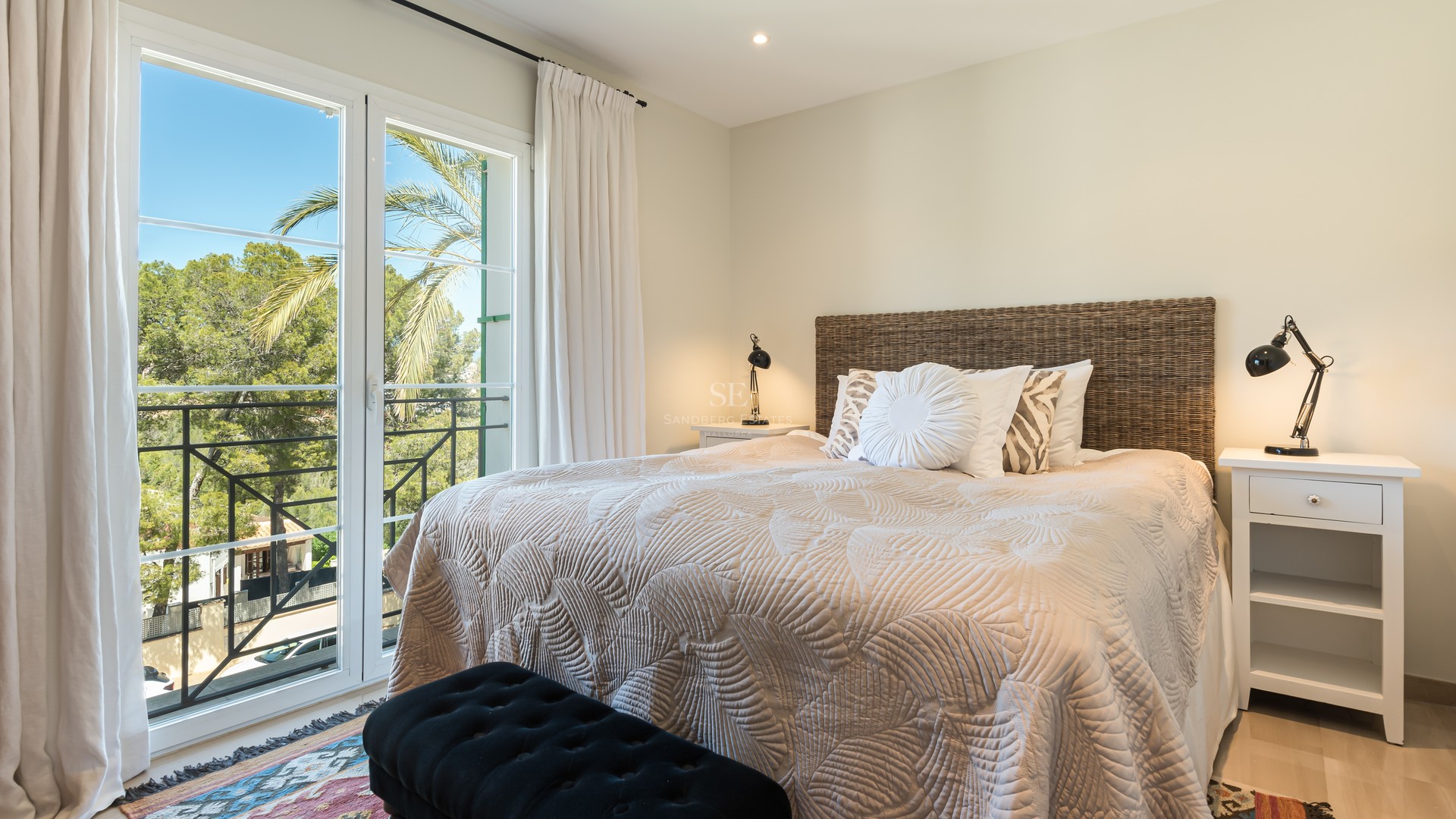 Bright bedroom featuring a woven wicker headboard, beige quilted bedding, and large French doors overlooking palm trees.
