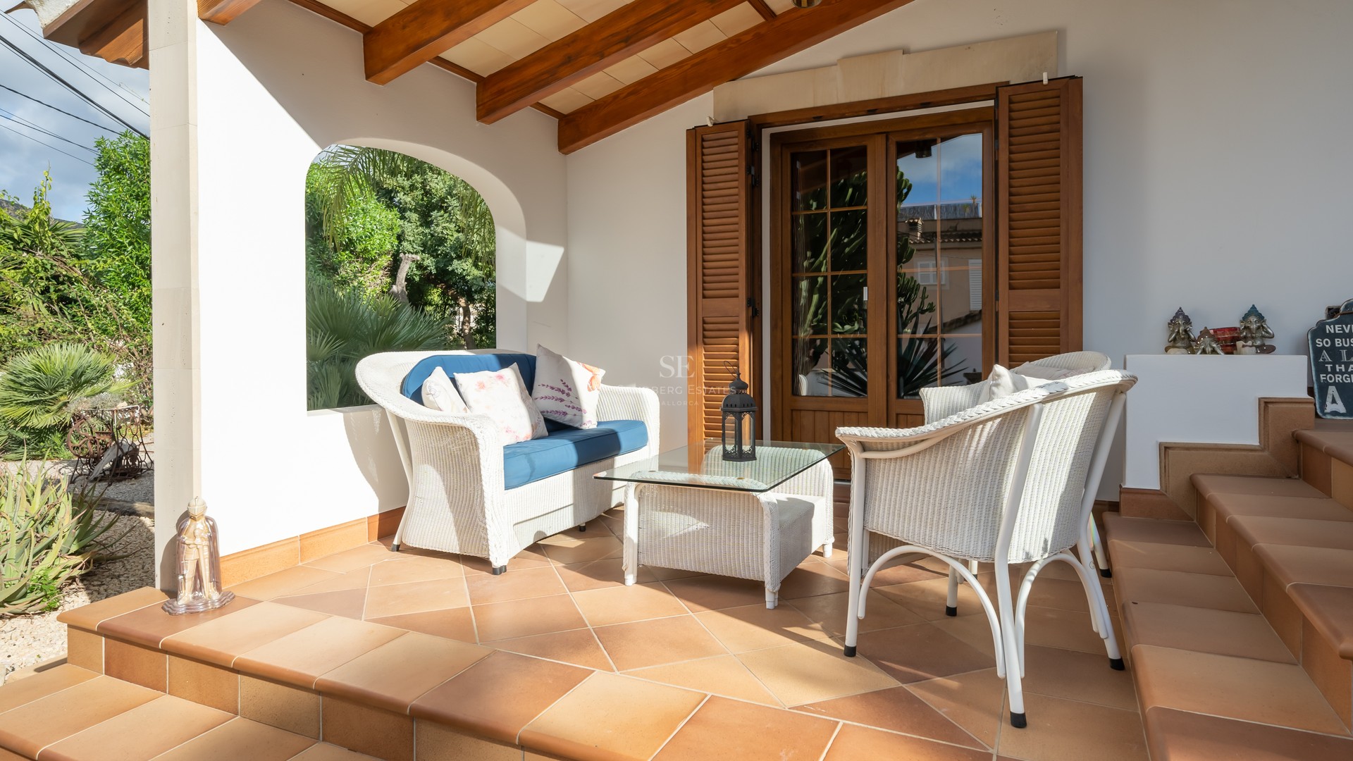 White wicker furniture on a terracotta-tiled terrace with exposed wooden beams and an arched view of a garden.