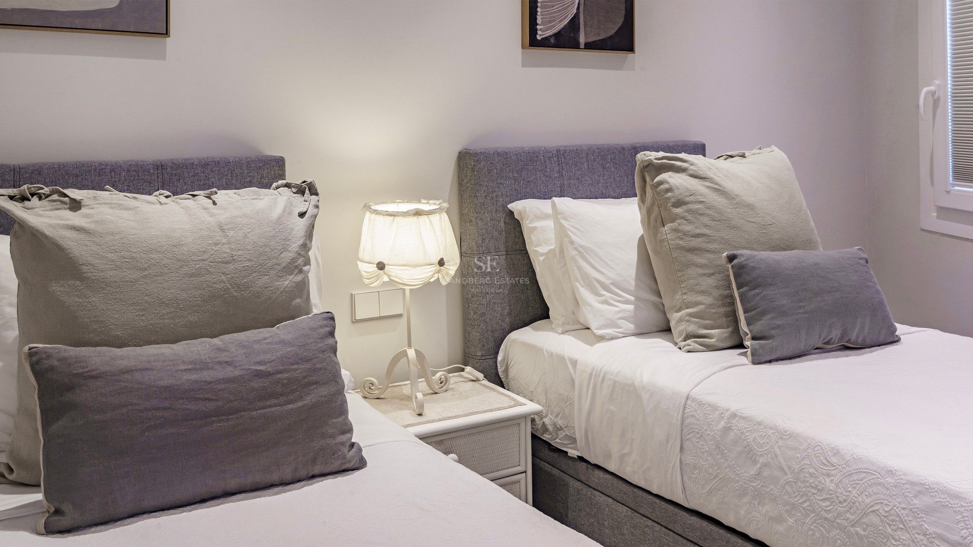 Two twin beds with grey upholstered headboards, white linens, and a lamp on a bedside table between them.