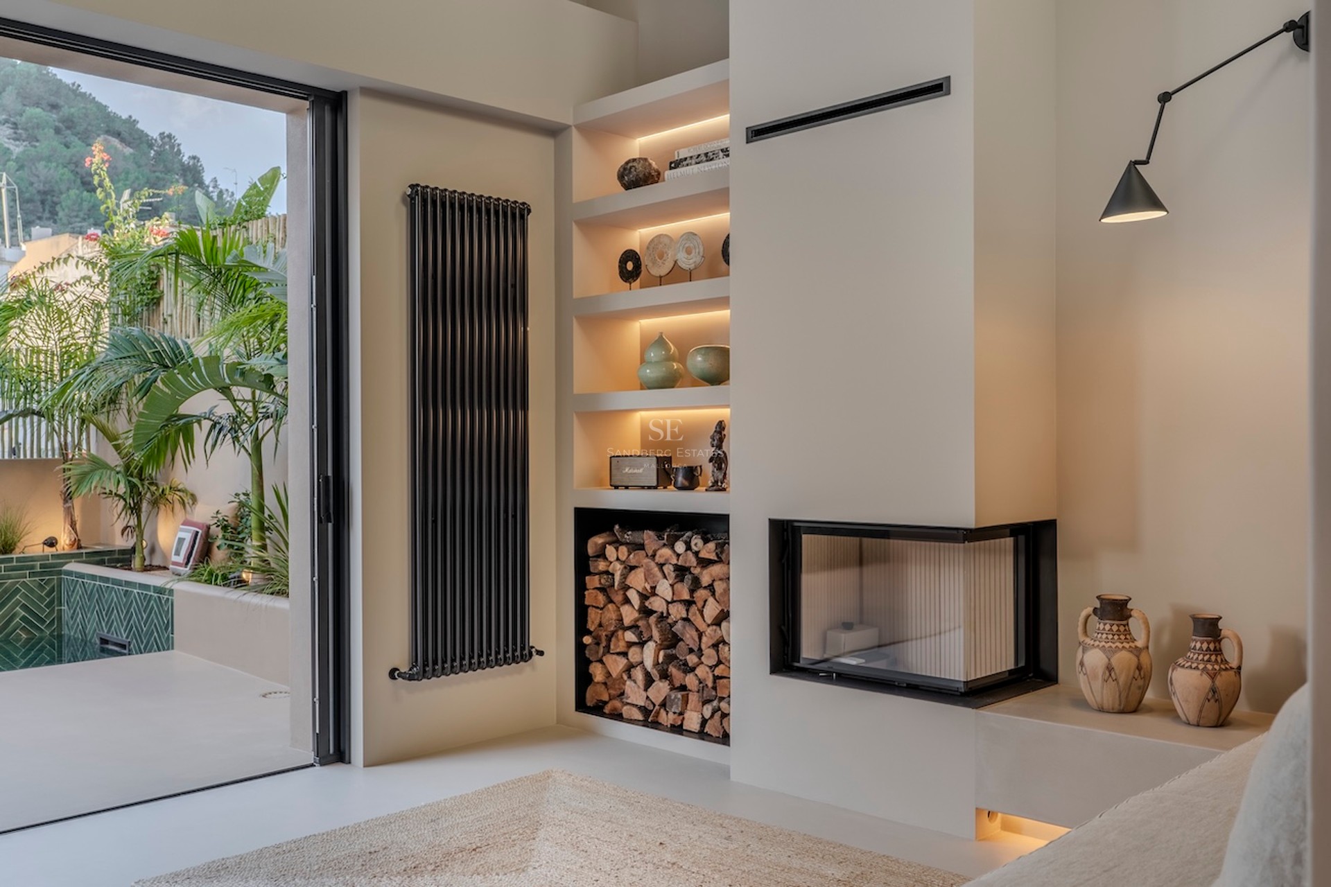 Espace de vie intérieur avec cheminée d'angle moderne, rangement à bois et vue sur une terrasse extérieure luxuriante.