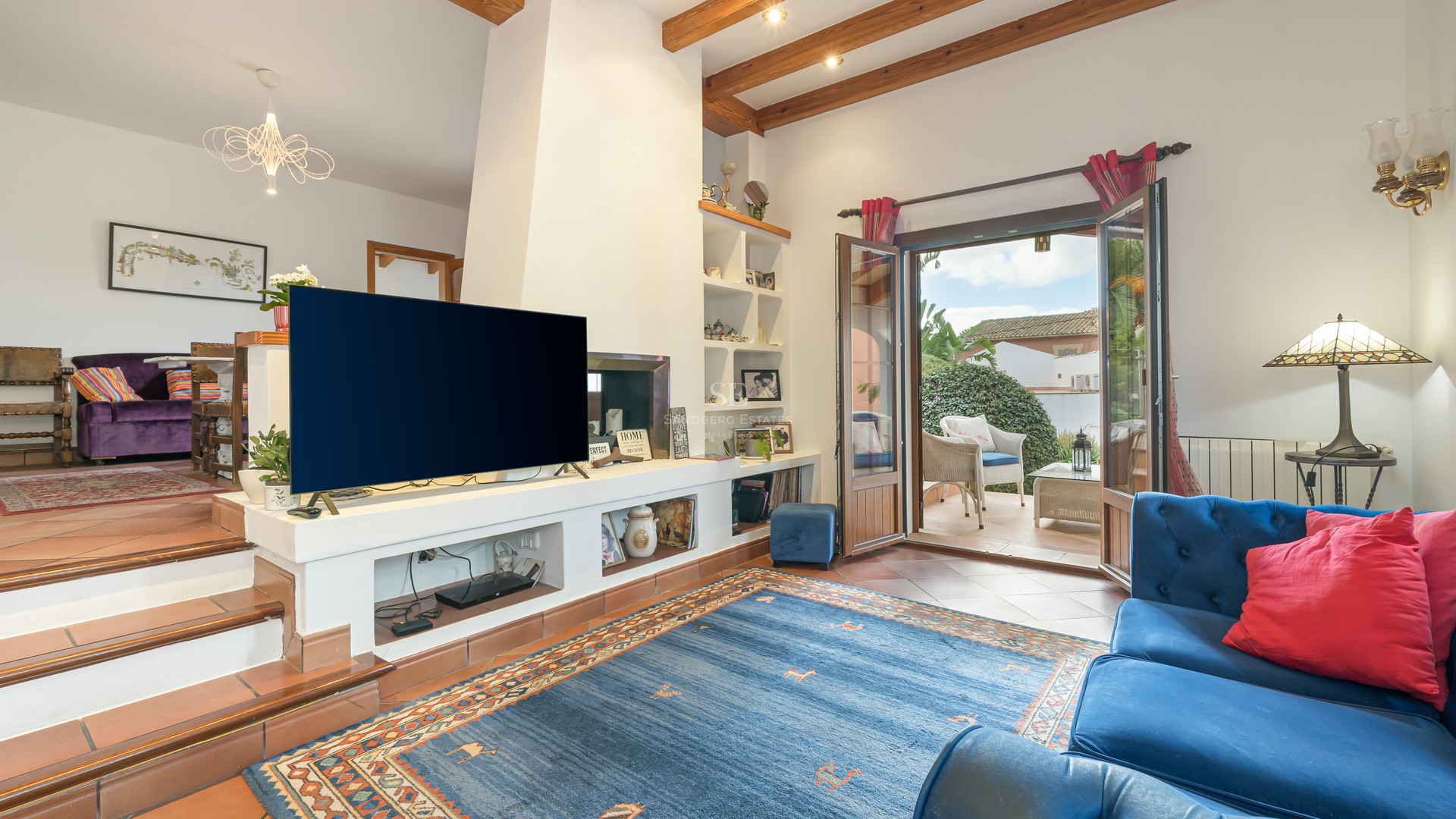 Living room featuring terracotta floors, exposed wooden beams, and open French doors leading to a sunlit terrace.