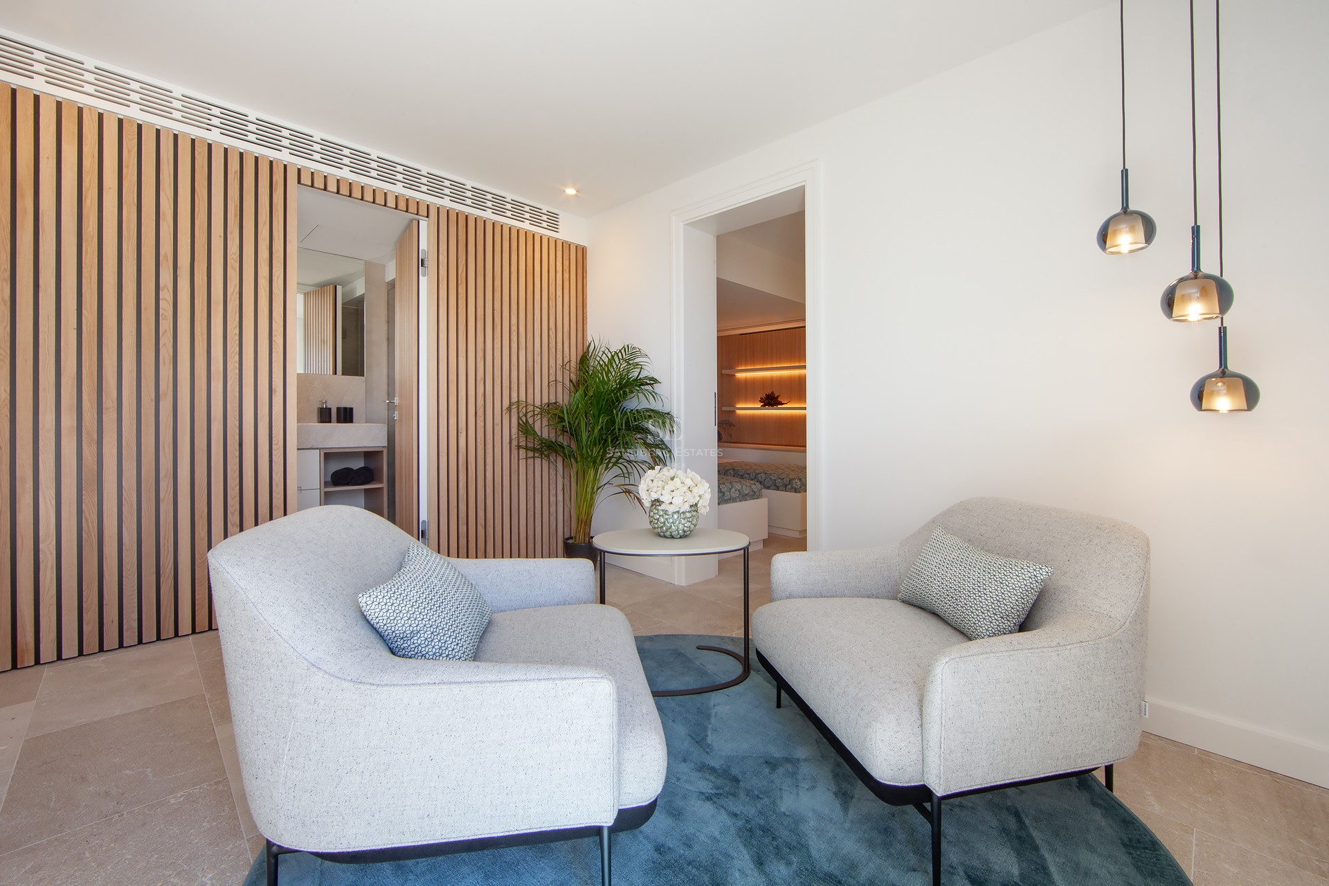 Suite parentale avec deux fauteuils gris, tapis bleu, mur en lattes de bois et vue sur la chambre.