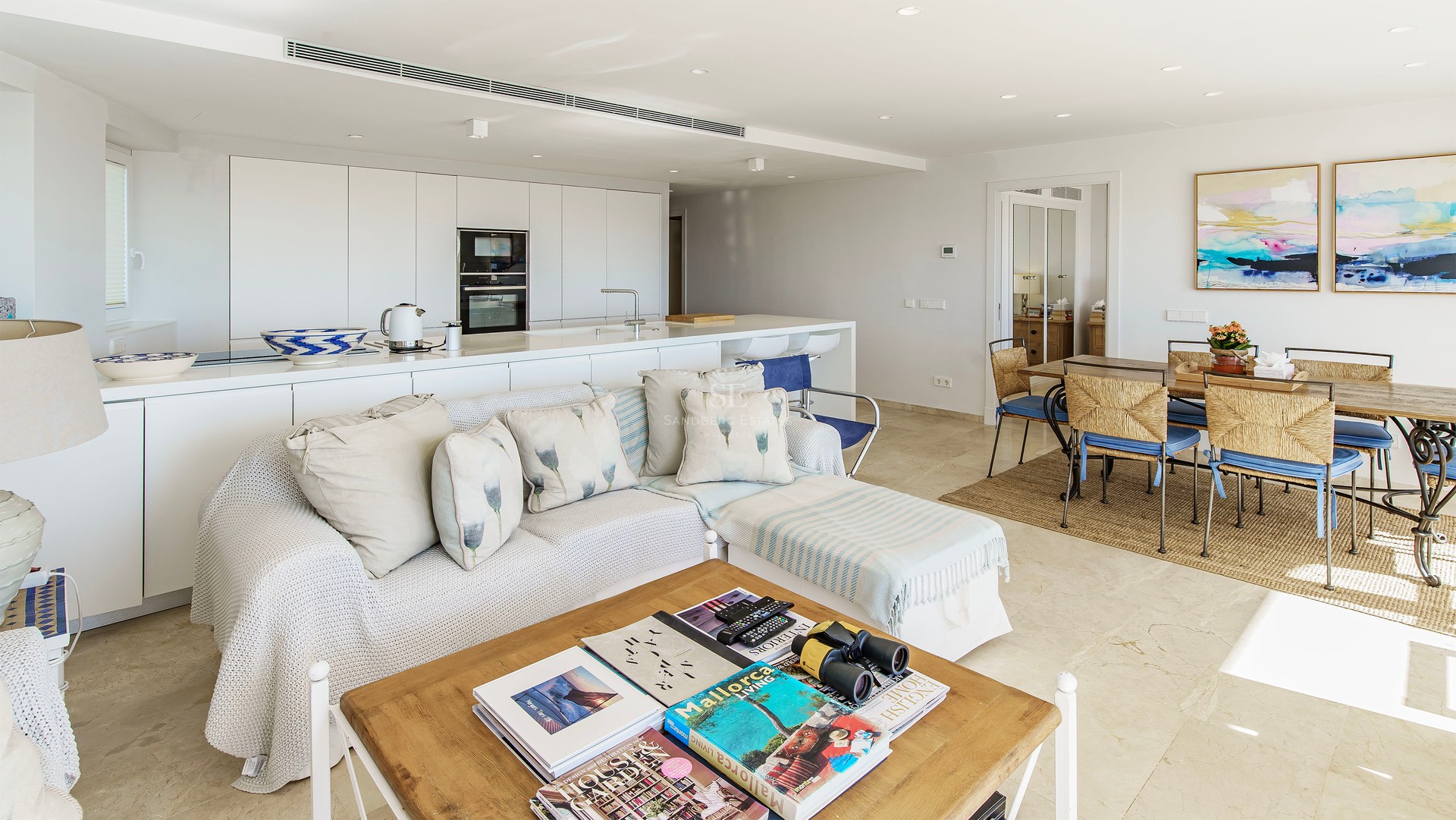 Bright open-plan living room with white sofa, wooden coffee table, dining set, and minimalist kitchen in the background.