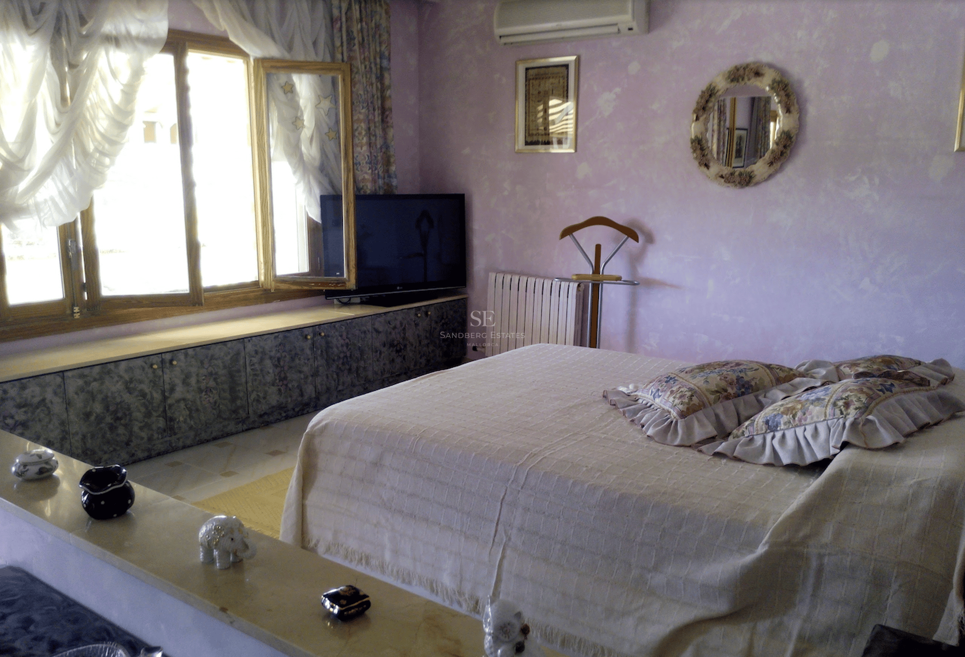 Dormitorio con paredes de color lila, cama grande con cojines florales y amplios ventanales de madera bajo luz natural.