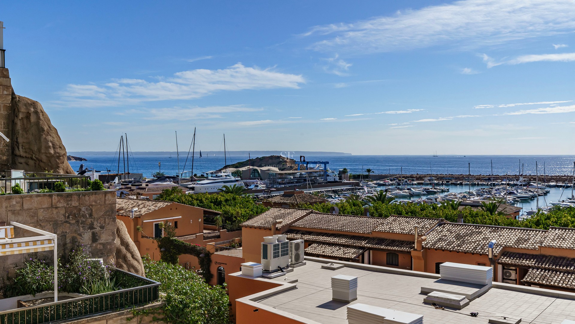 Elevated view of a Mediterranean marina with terracotta roofs, white yachts, and the blue sea under a clear sky.