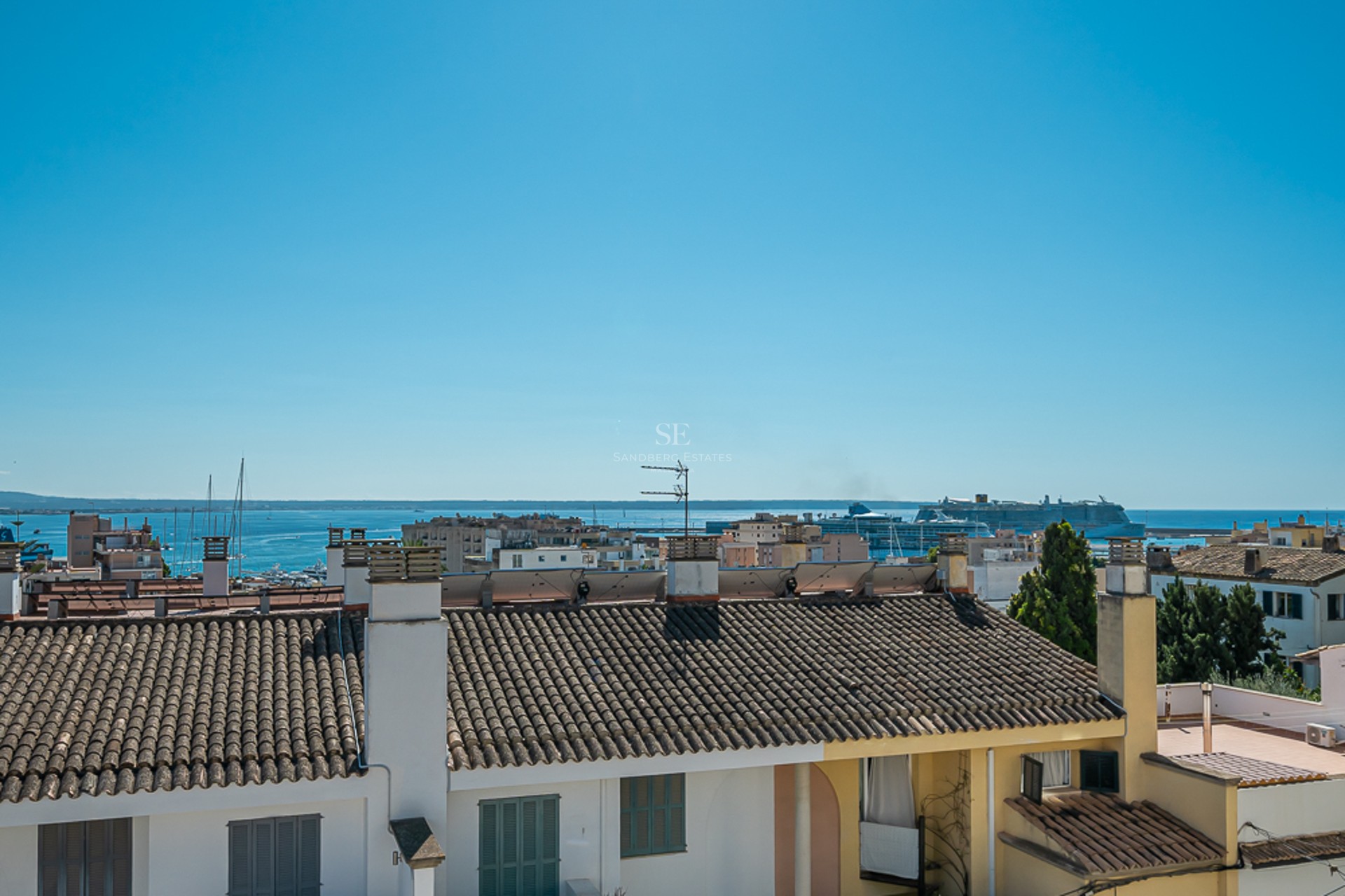 Elevated view over terracotta rooftops towards a blue sea with a large cruise ship in the harbor under a clear sky.