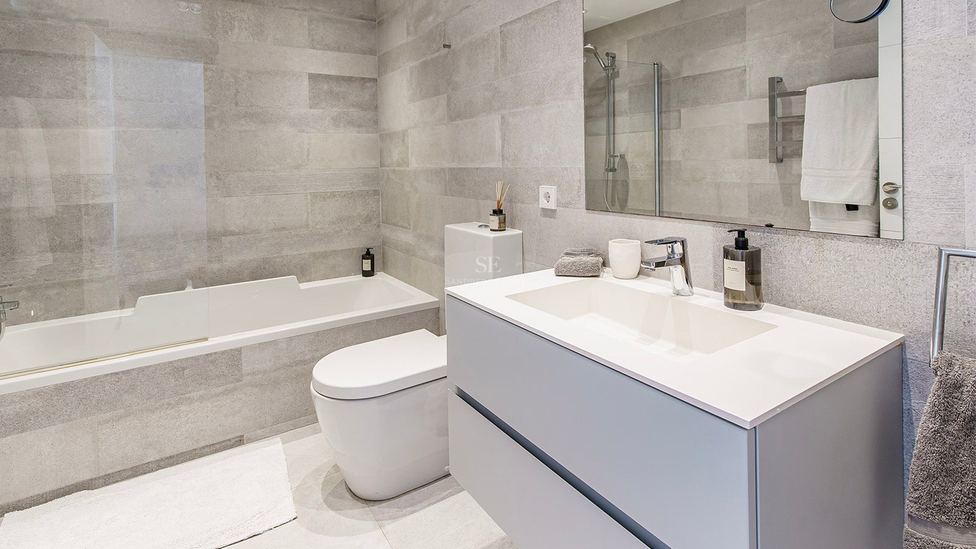 Contemporary bathroom featuring grey stone-effect tiles, a white bathtub, toilet, and modern vanity with a large mirror.