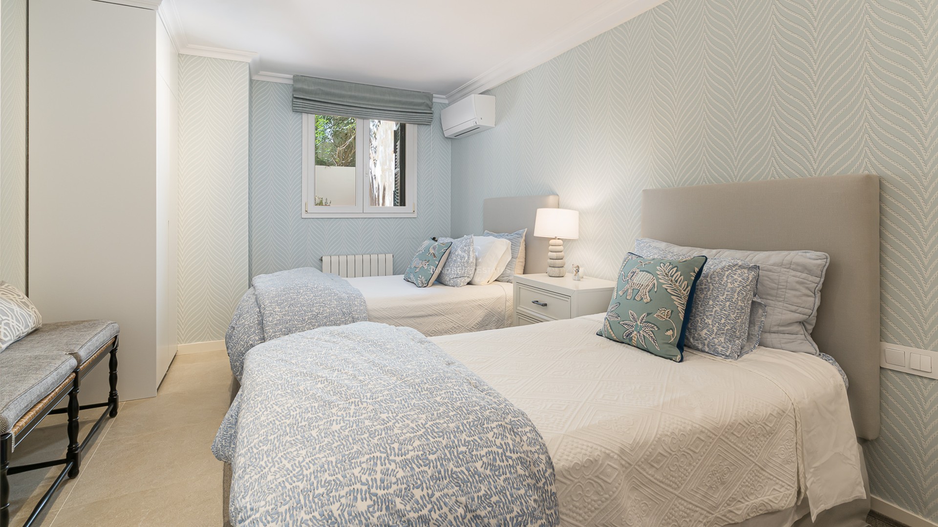 Bright room with two single beds, light blue patterned wallpaper, built-in wardrobes, and stone flooring.