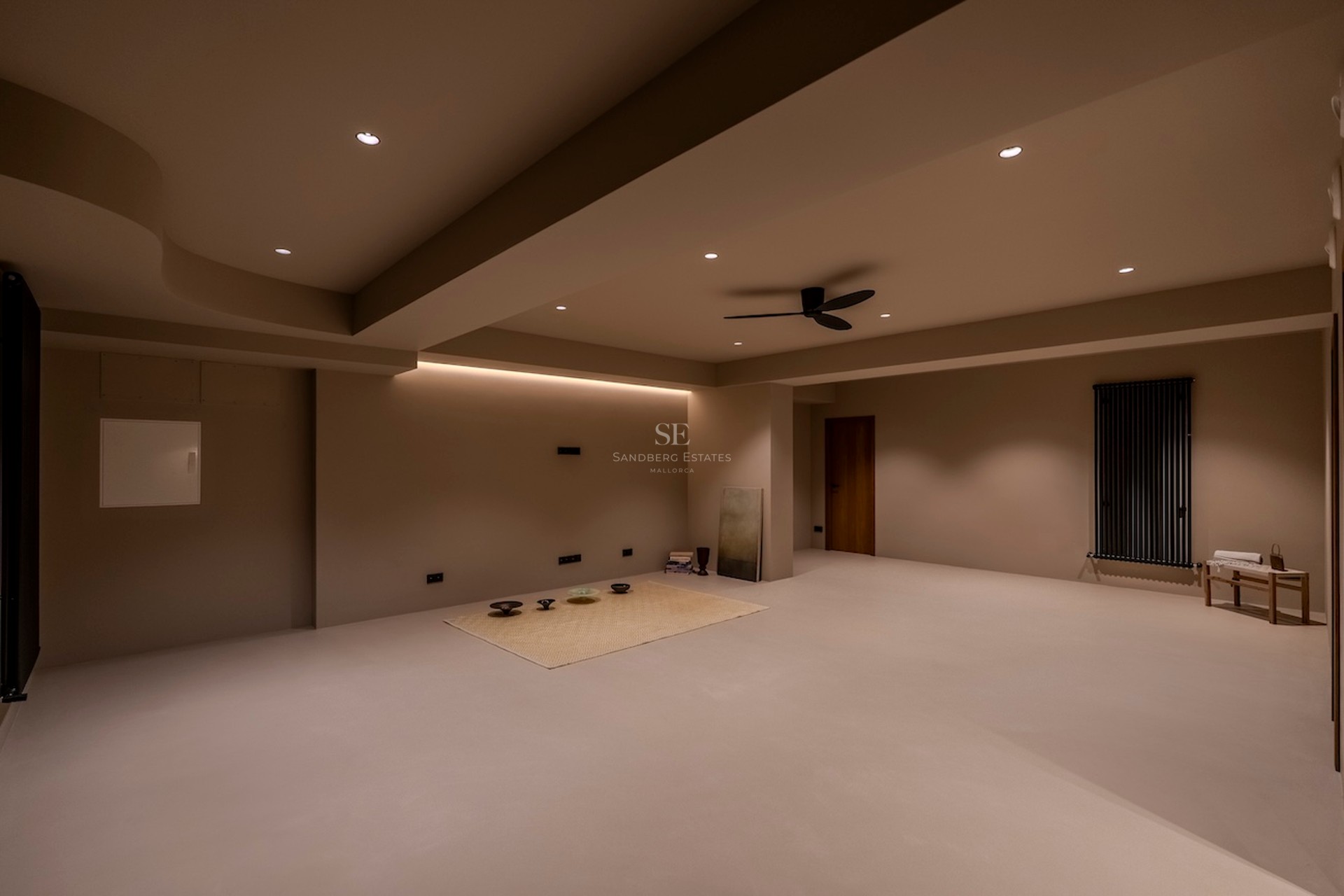 Une grande pièce minimaliste aux murs et sols beiges, avec éclairage encastré et ventilateur de plafond.