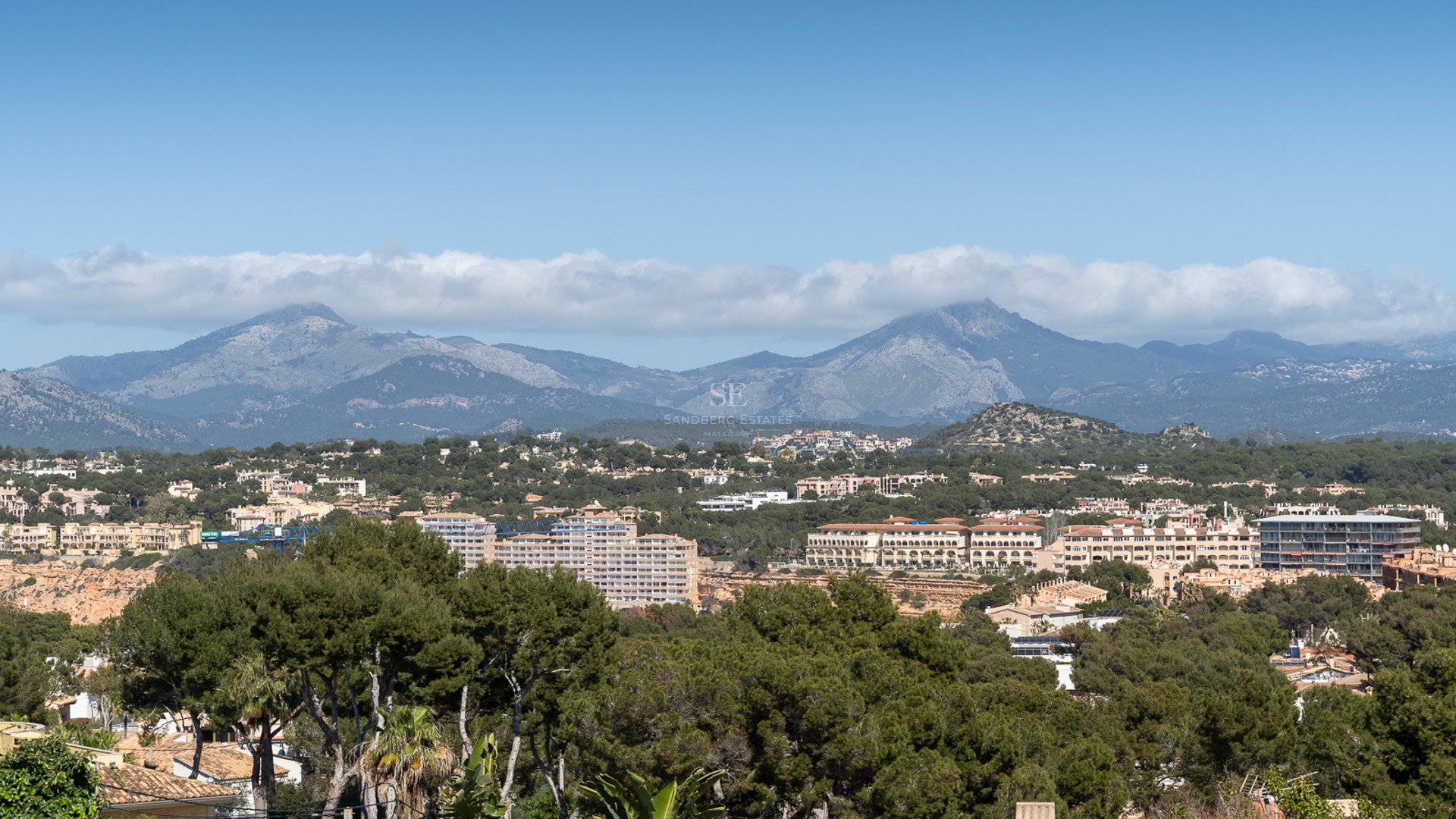 Flygvy över medelhavslandskap med tallskogar, bostadshus och berg under en klarblå himmel.