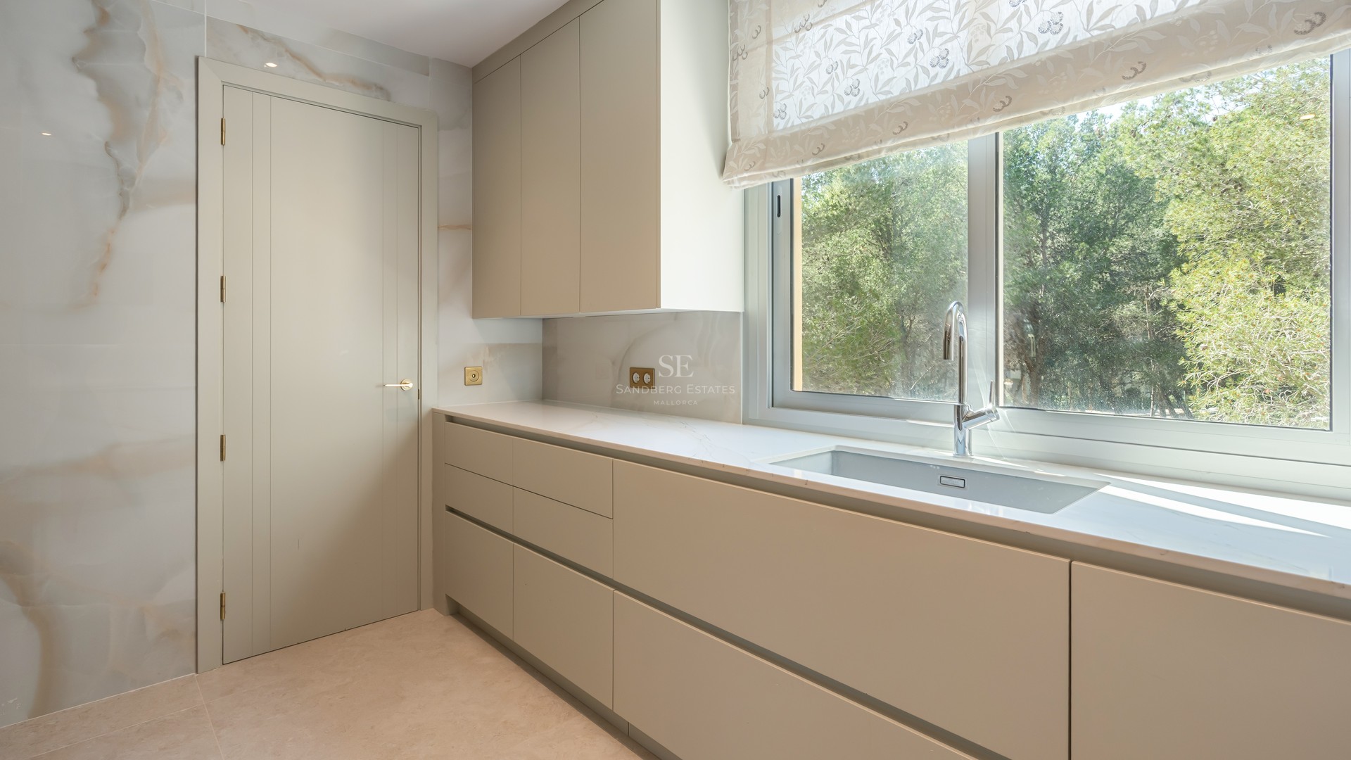 Minimalist kitchen featuring cream handleless cabinets, a large window overlooking greenery, and a stone backsplash.