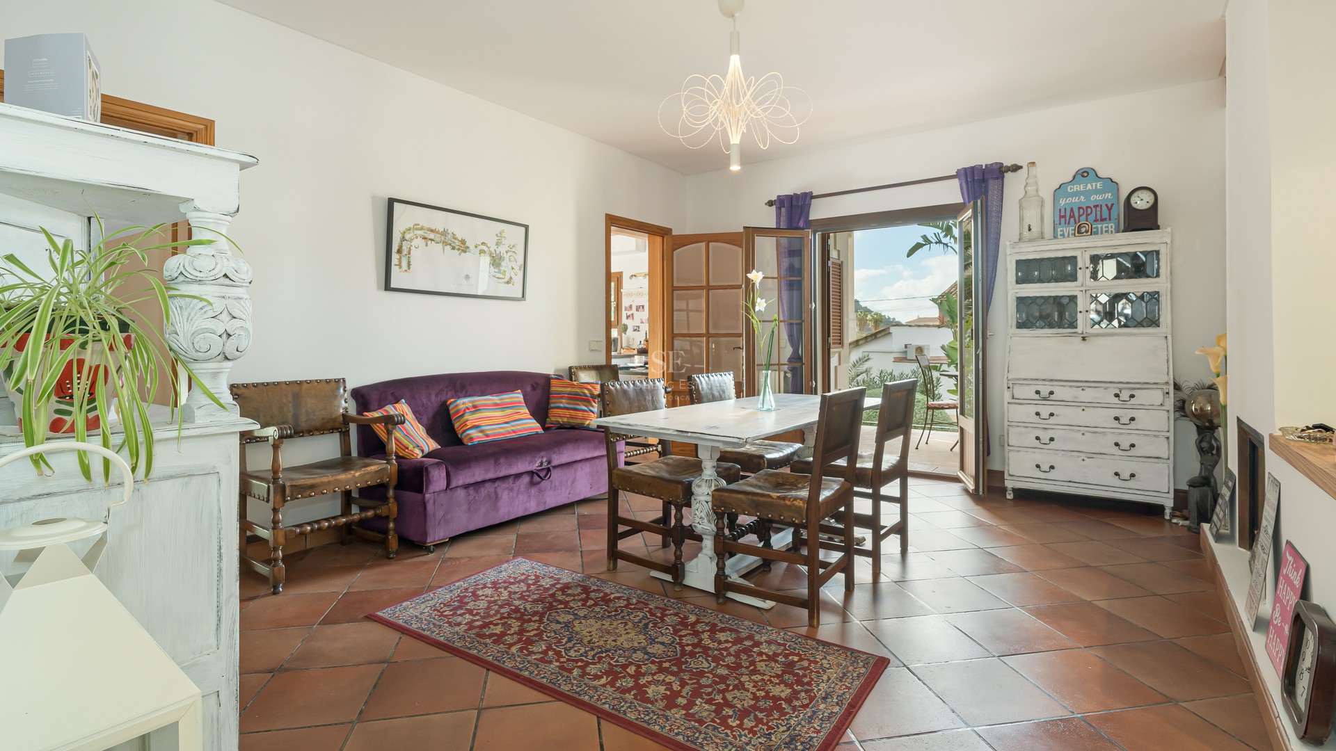 Dining room with terracotta floors, purple velvet sofa, and white wooden table opening to a sunny balcony.
