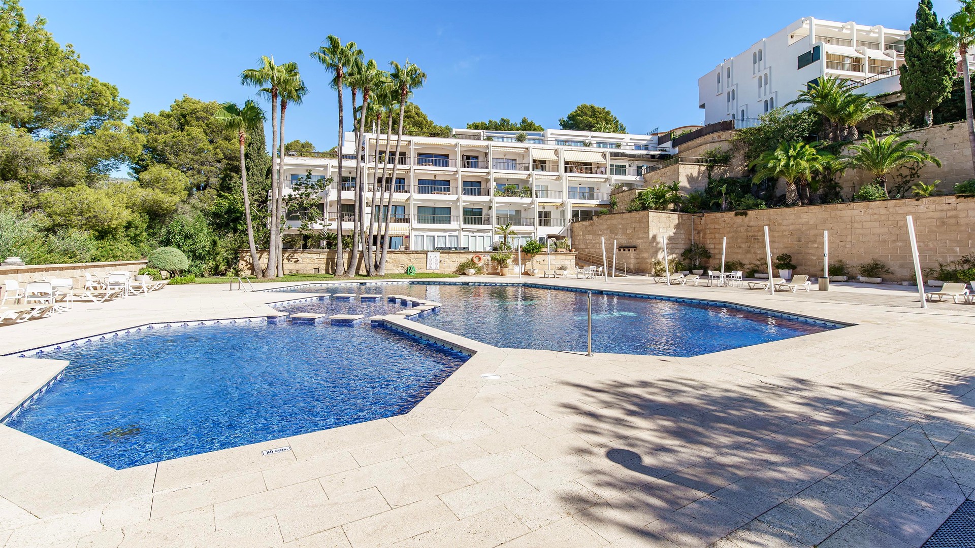 Large blue swimming pool surrounded by stone paving, palm trees, and white apartment buildings under a clear sky.