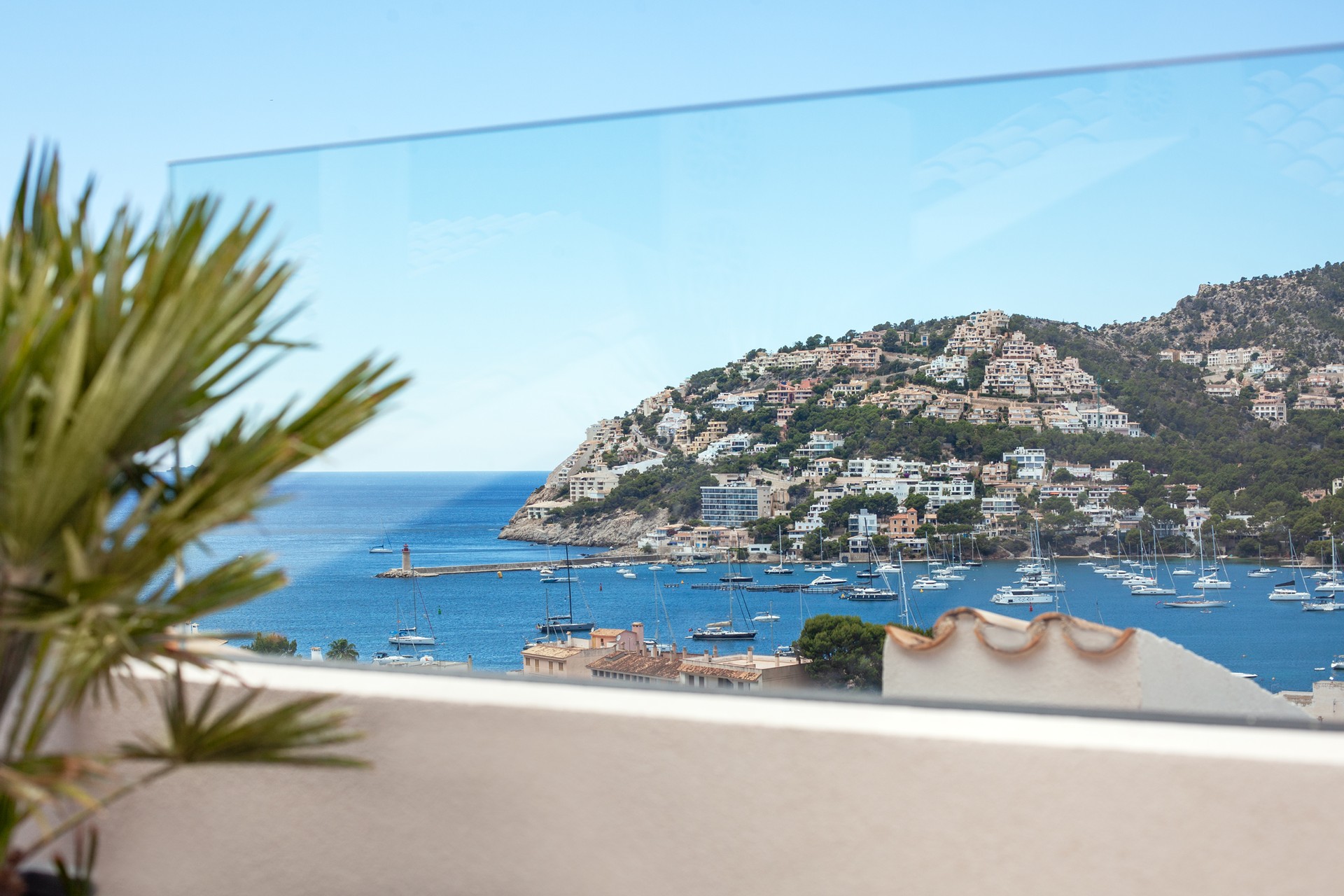 Vue sur un port méditerranéen avec bateaux et villas à flanc de colline à travers un garde-corps en verre.