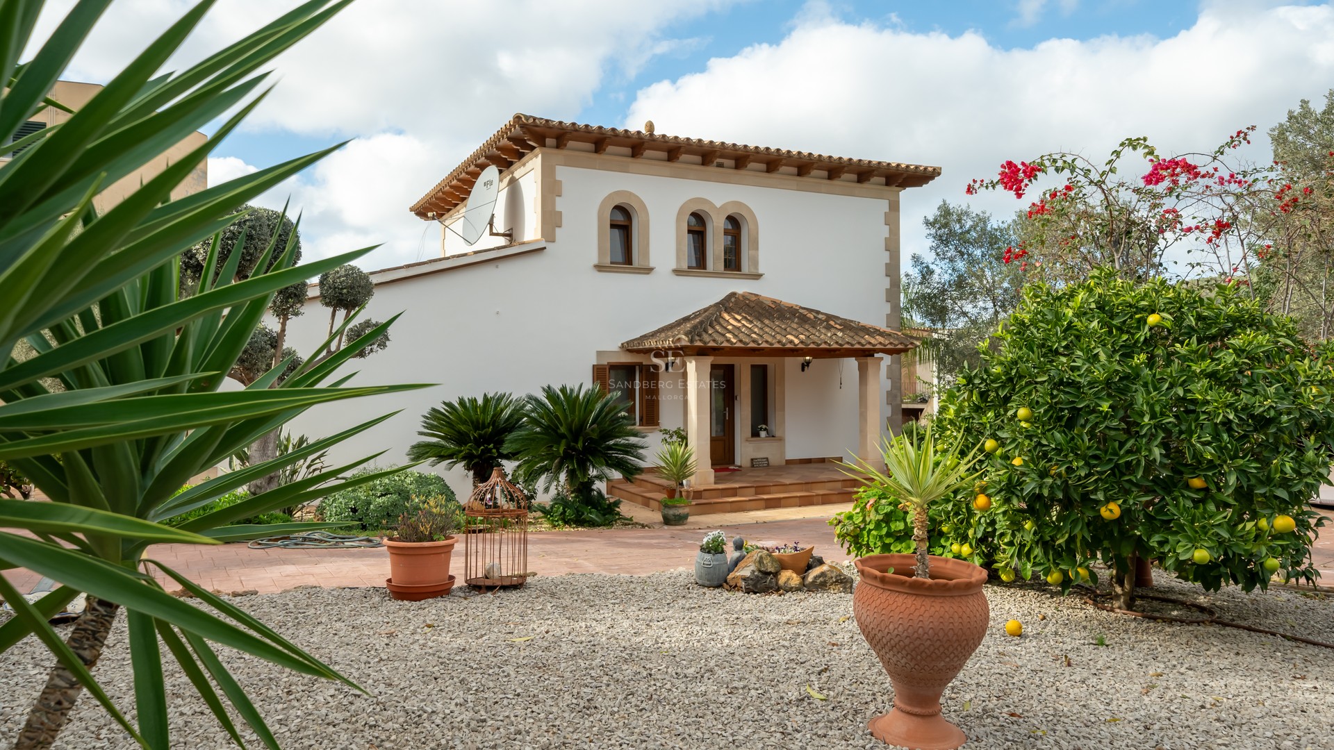 White two-story villa with terracotta roof, arched windows, and a landscaped garden featuring lemon trees and gravel.