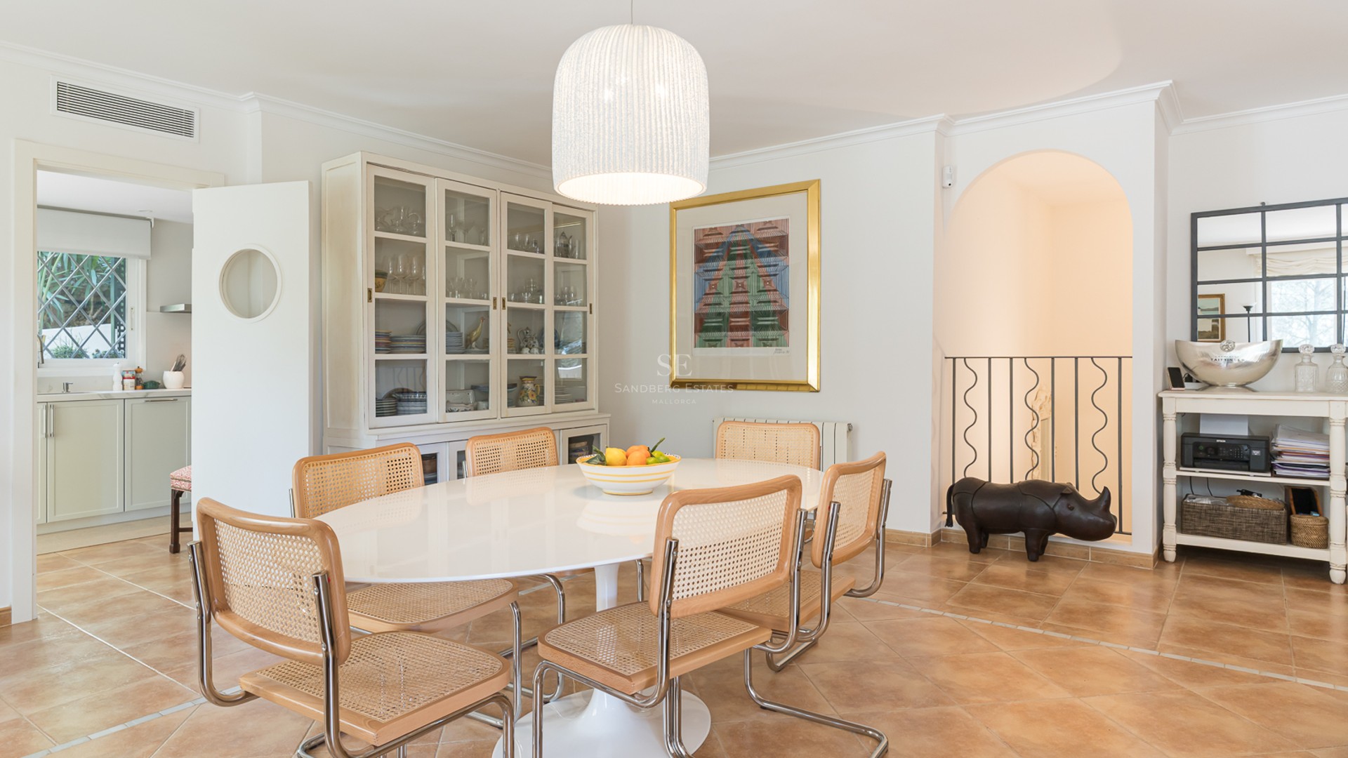 Bright dining room featuring an oval white table, rattan chairs, terracotta floors, and a large display cabinet.