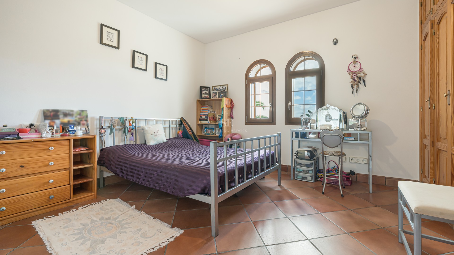 Bedroom featuring terracotta tiles, metal bed frame, arched wooden windows, and built-in wardrobes under natural light.
