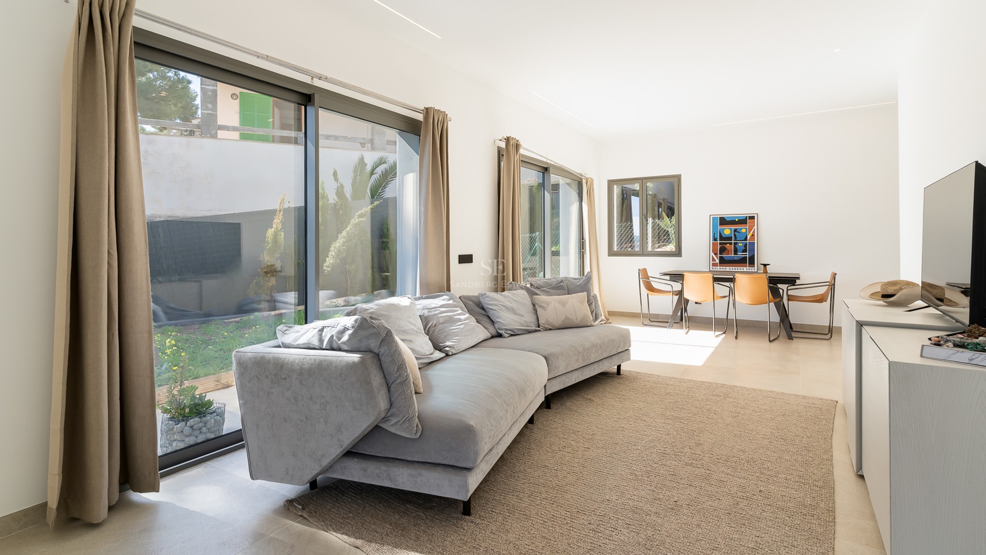 Ljust vardagsrum med grå soffa, stora glaspartier mot trädgården och matplats med läderstolar.