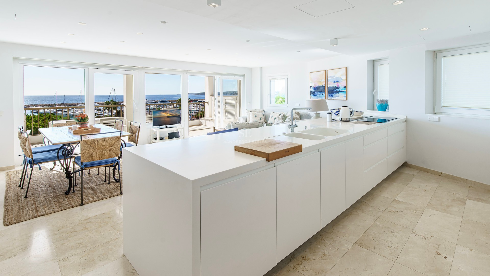 Minimalist white kitchen island leading to a dining area with a panoramic view of the harbor and sea.