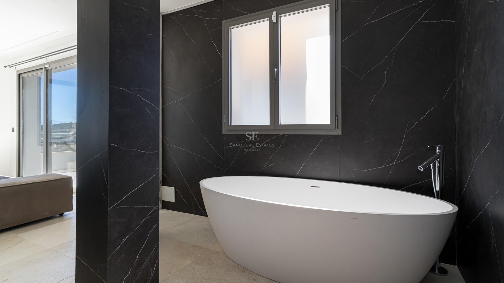 Ett modernt badrum med ett vitt ovalt fristående badkar mot en svart marmorvägg och ett frostat fönster.