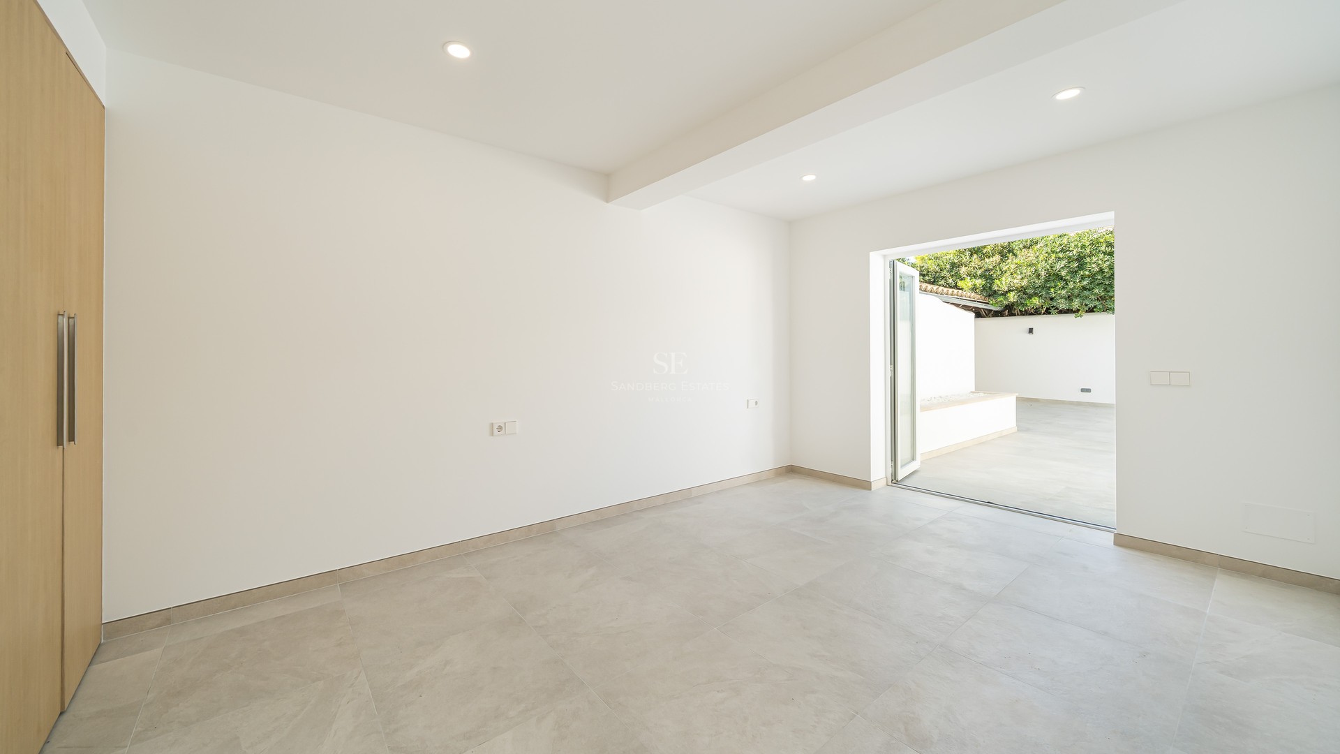 Pièce lumineuse et vide avec sol gris, murs blancs, placard en bois et porte vitrée donnant sur un patio.