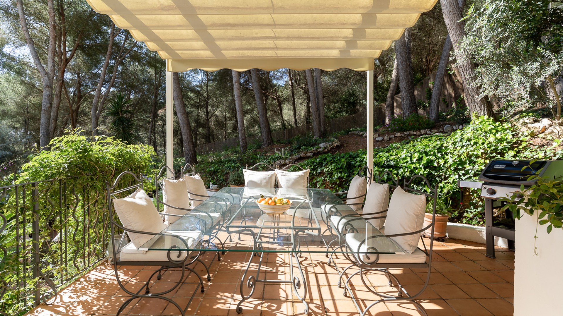 Covered terrace with wrought iron dining table, glass top, and cream cushions, surrounded by lush pine trees.