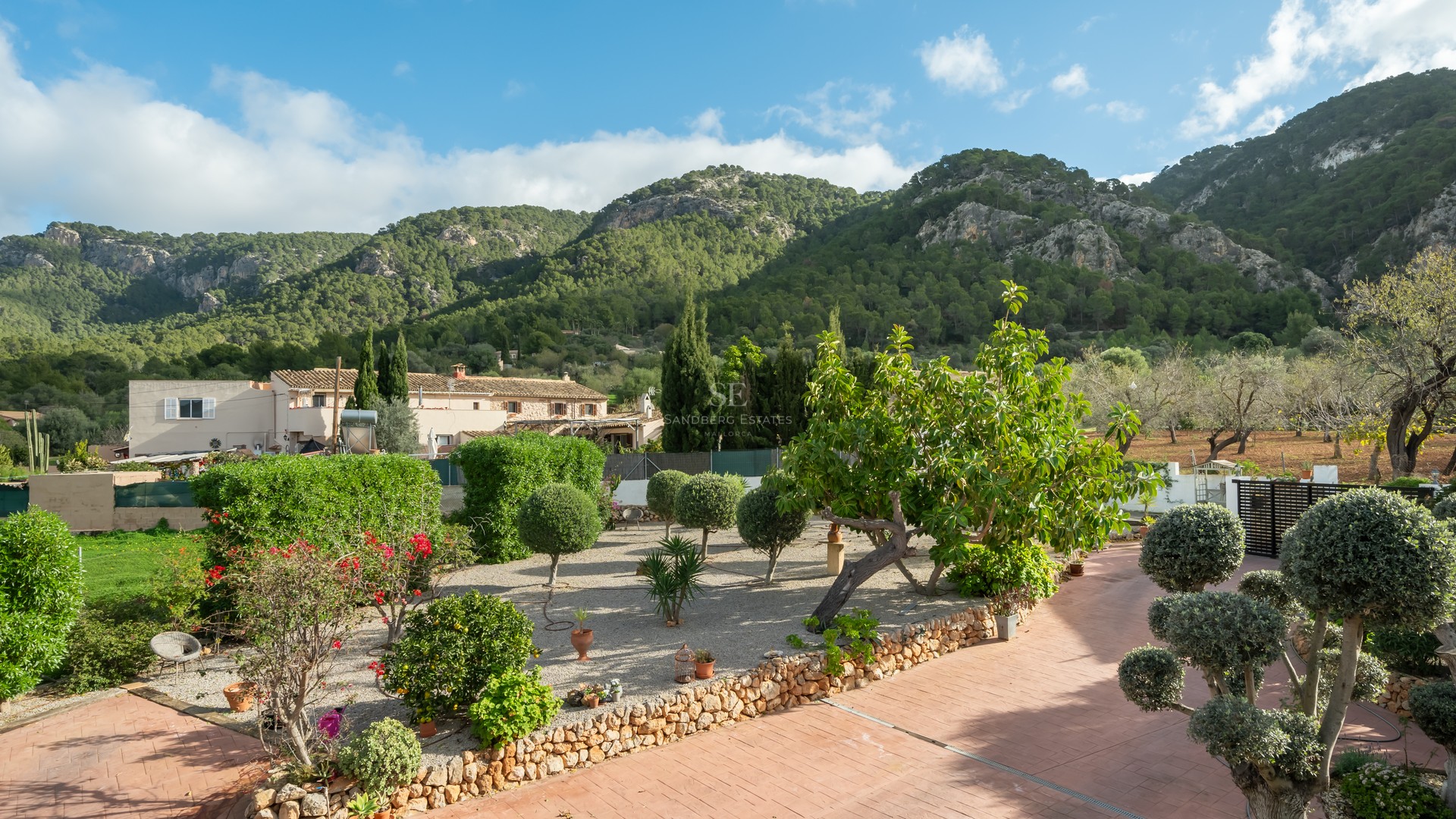 Landscaped garden with terracotta paths, gravel, and manicured shrubs against a backdrop of pine-covered mountains.