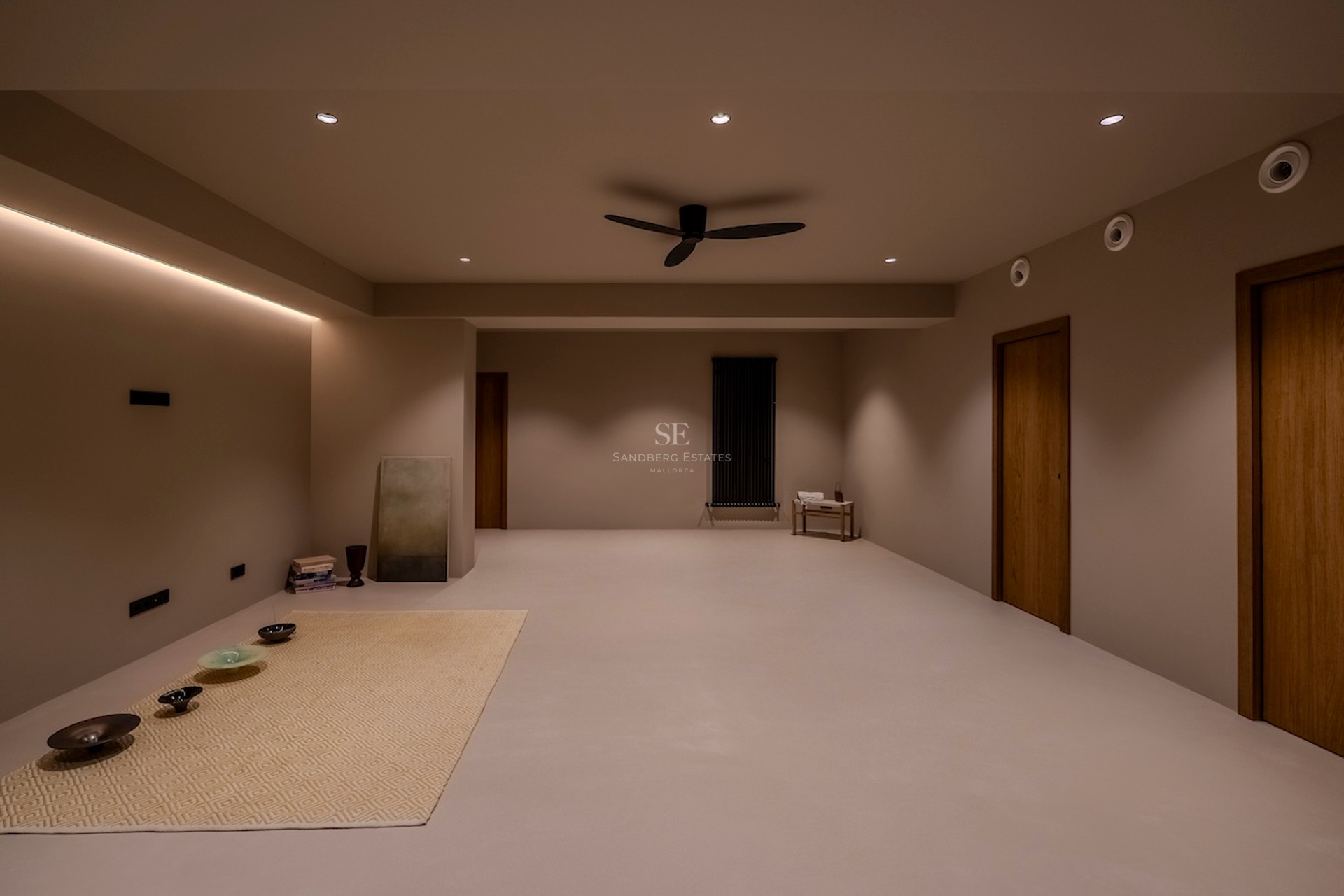 Une grande pièce minimaliste avec un éclairage chaleureux, des murs taupe, des portes en bois et un ventilateur noir.