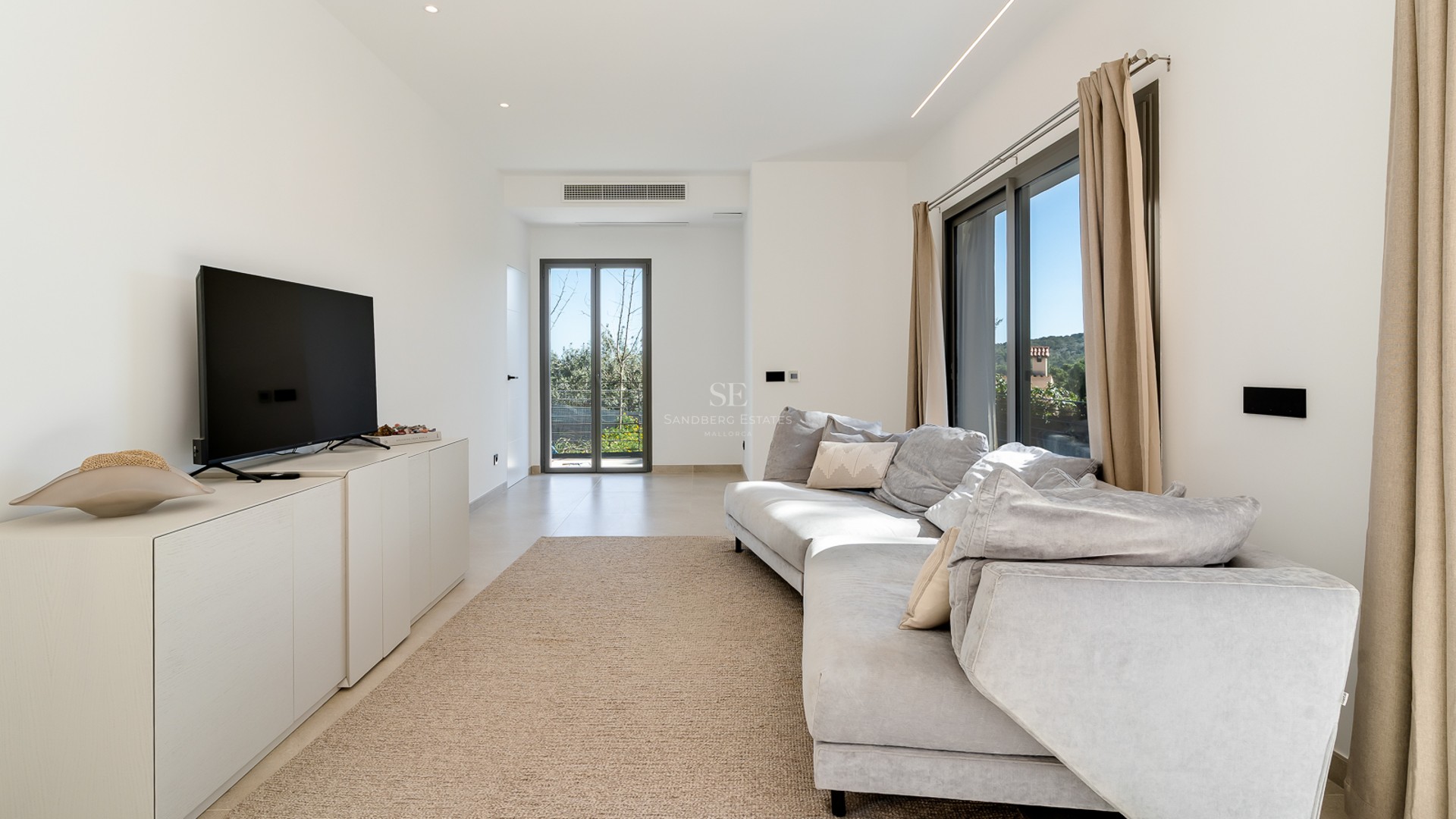 Modernt vardagsrum med grå soffa, naturfibermatta och stora glaspartier som släpper in rikligt med dagsljus.