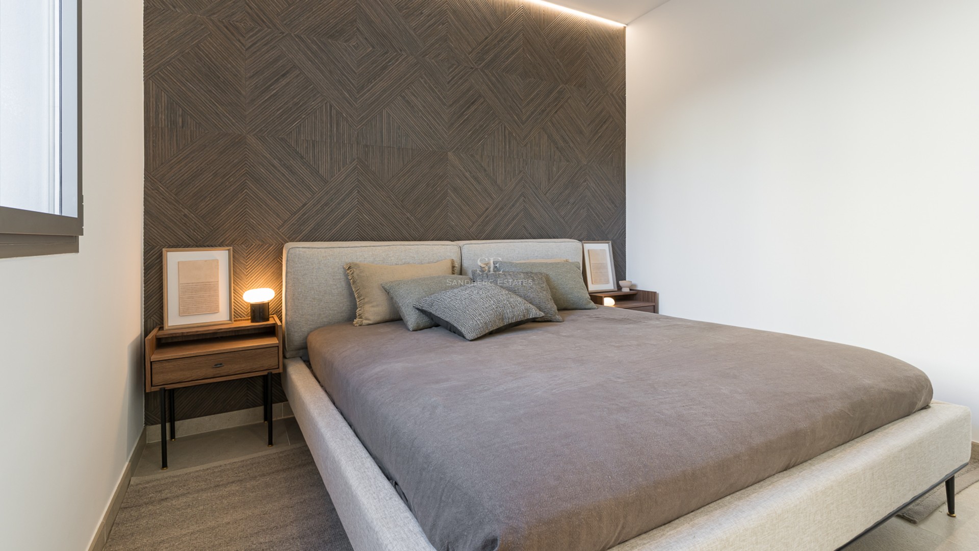 Ett modernt sovrum med en geometrisk fondvägg i trä, stoppad säng och nattduksbord i valnötsträ.